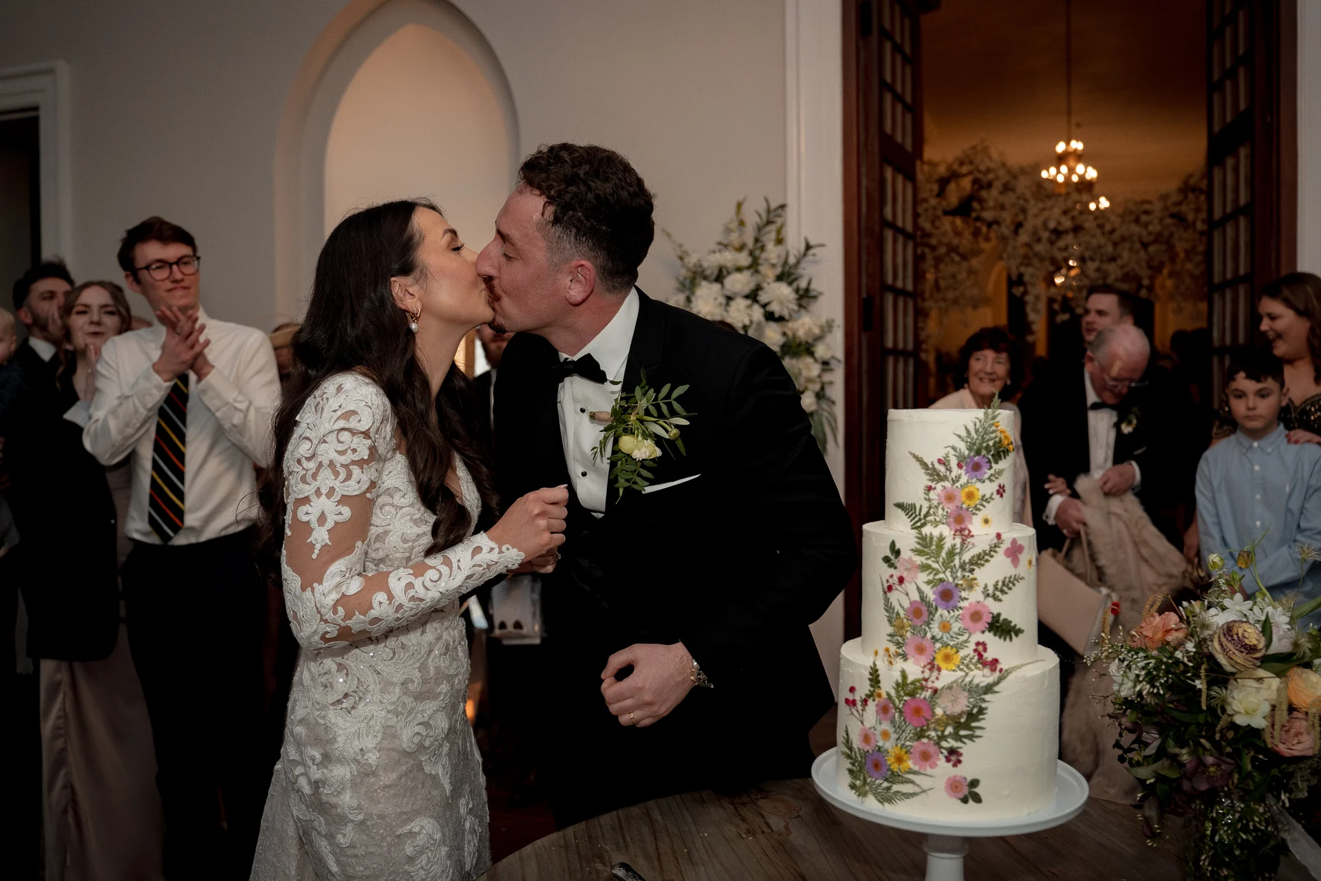 Newlyweds share a romantic kiss after cutting their tiered wedding cake adorned with pressed wildflowers, surrounded by cheering guests indoors.