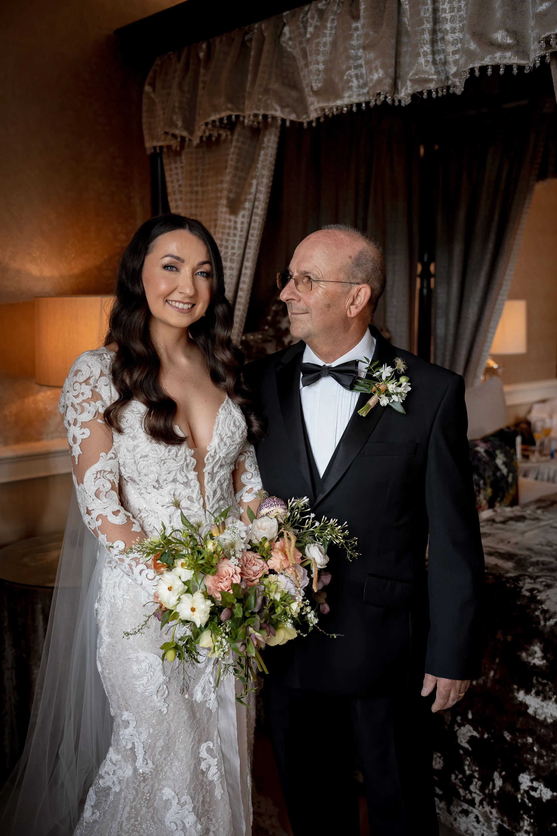 A smiling bride in a plunging lace long-sleeve gown stands proudly holding a hand-tied floral bouquet beside her father in a classic black tuxedo.