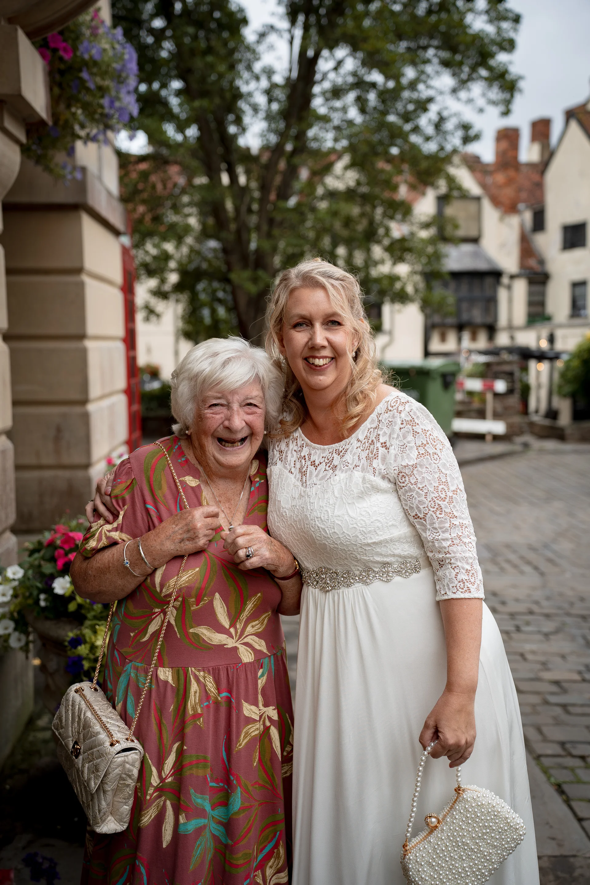 A mature bride in a lace gown and her grandmother share a joyful, laughing embrace outside on a cobbled courtyard.