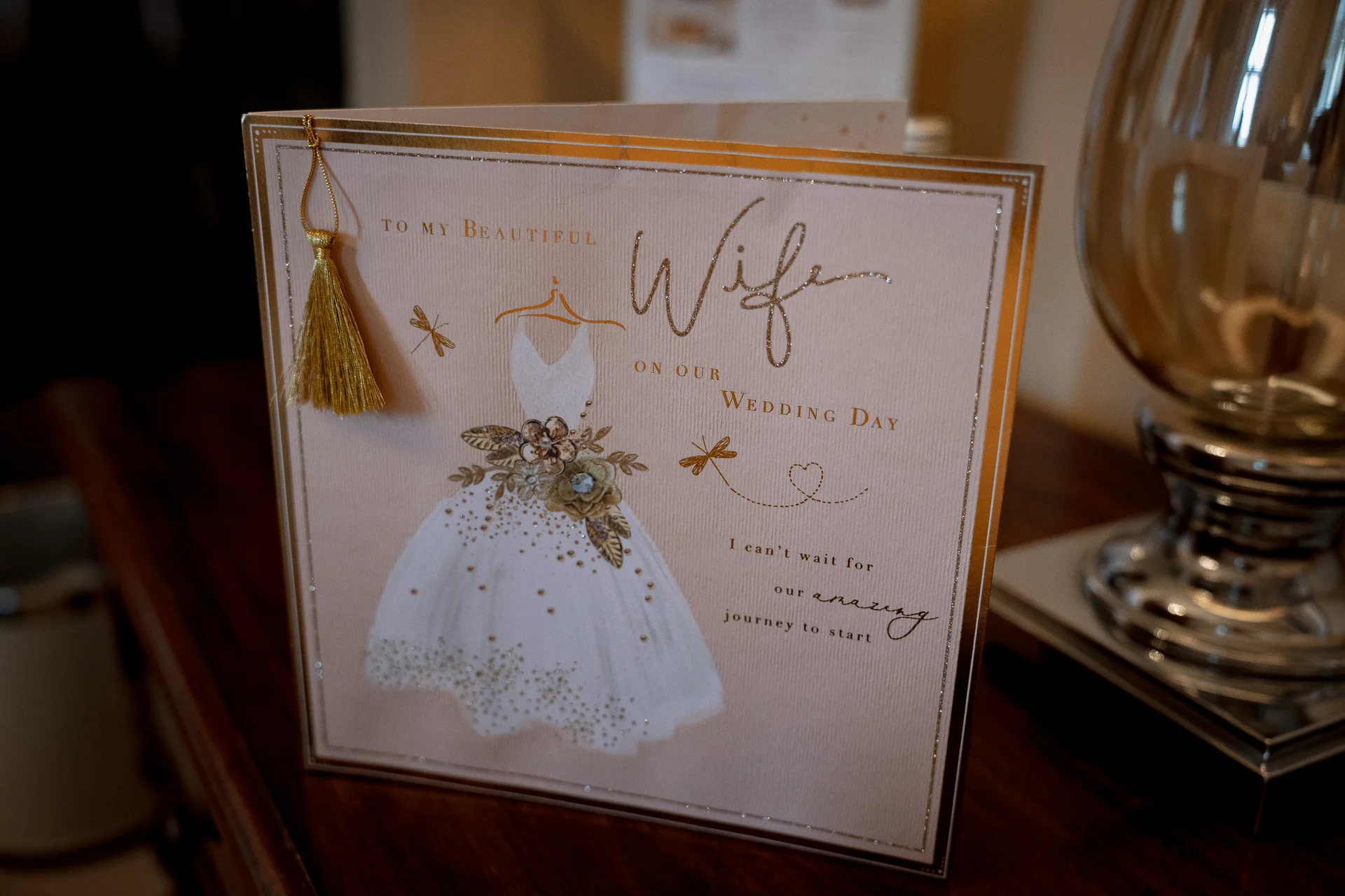 A greeting card addressed to a beautiful wife on her wedding day sits on a wooden surface next to a decorative glass lamp.