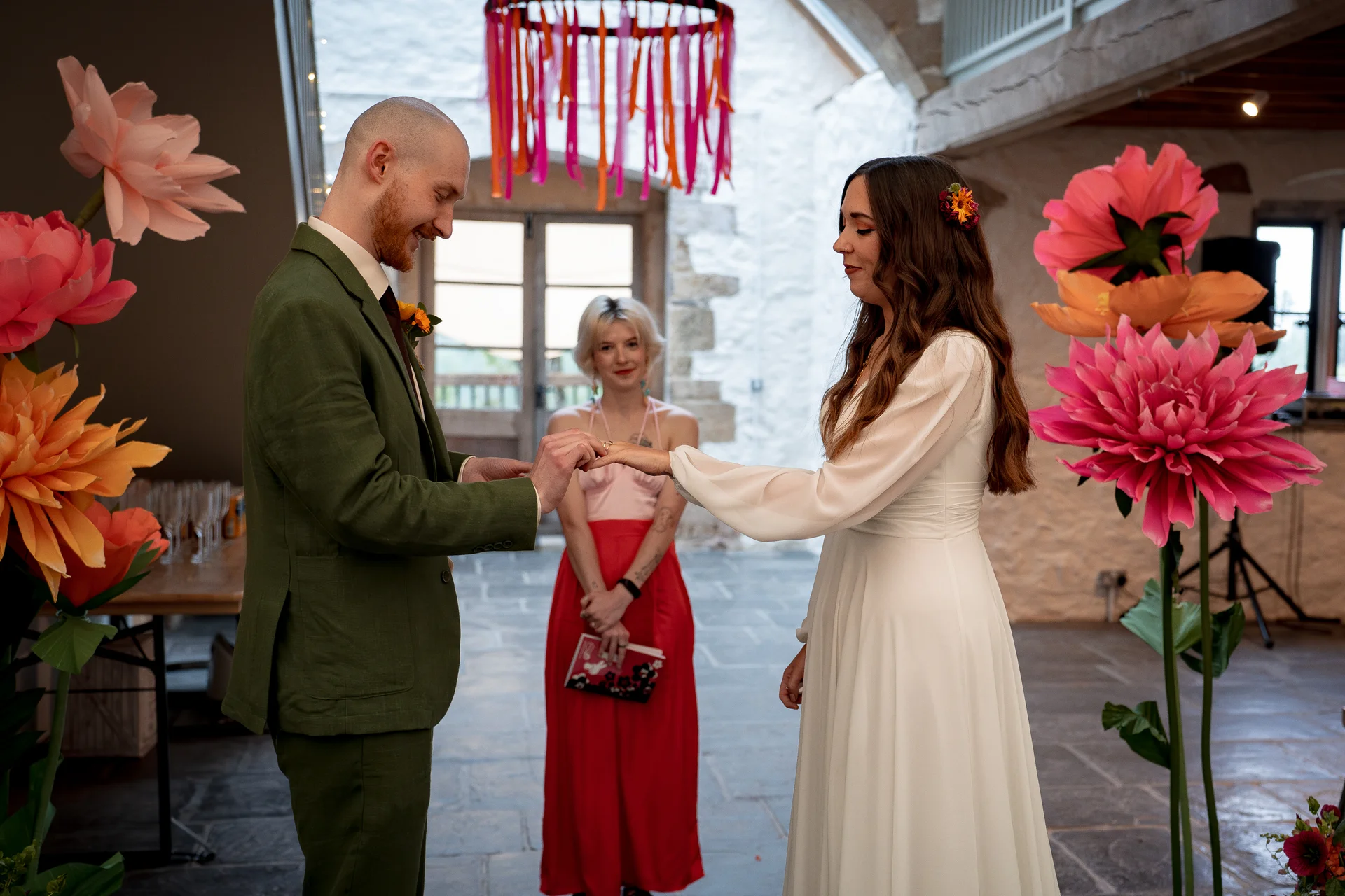 A groom in an olive green suit places a ring on the bride's finger during an indoor ceremony framed by giant paper flowers and a celebrant standing in the background.