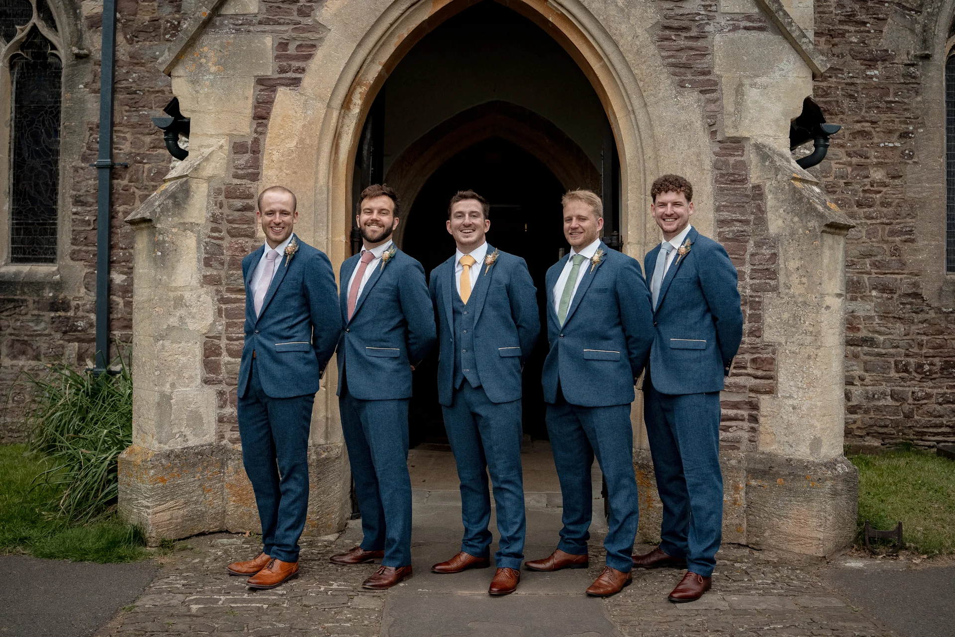 Five groomsmen in matching blue tweed suits and brown shoes pose together outside a historic stone church entrance.