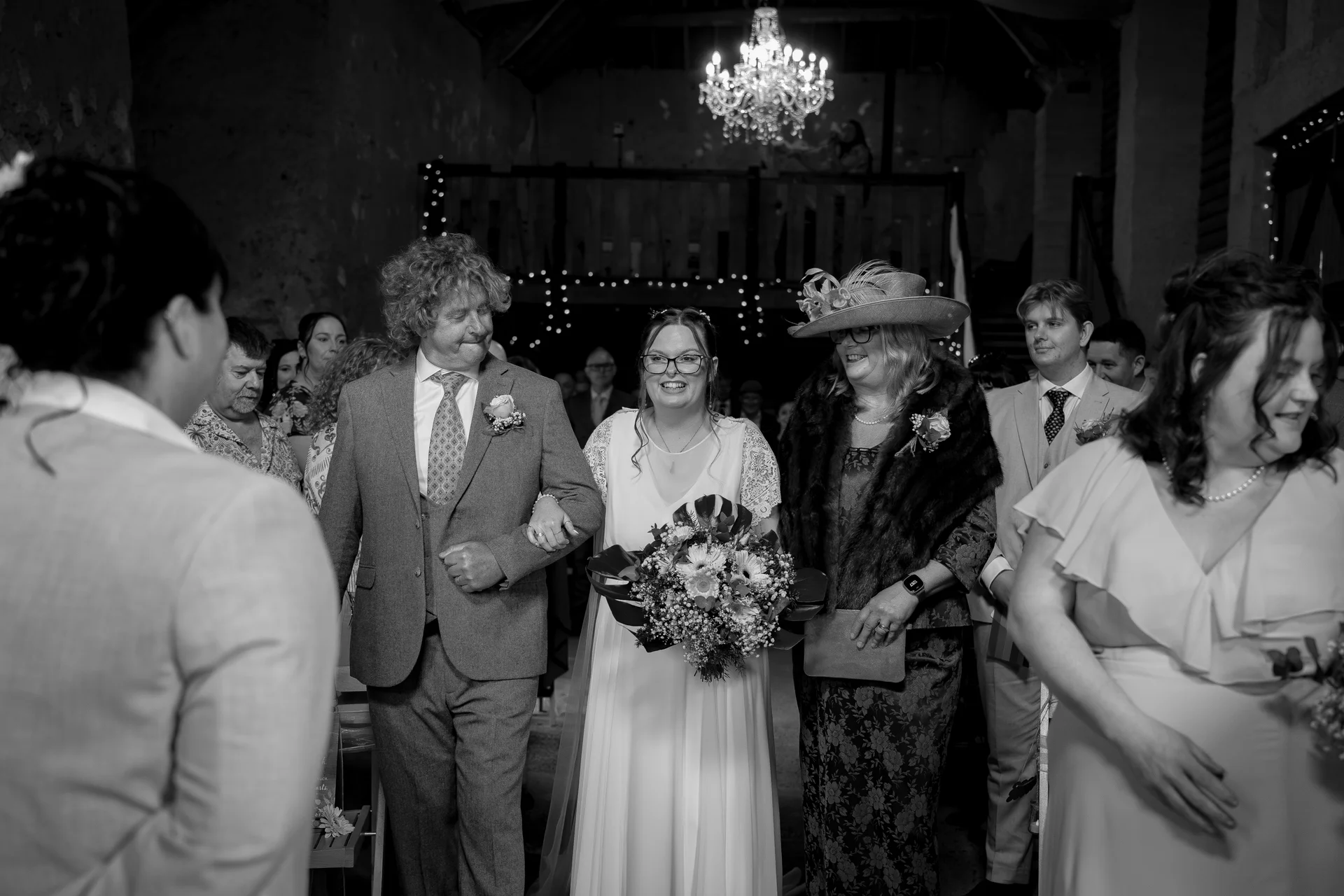 A smiling bride walks down the aisle arm in arm with her father, flanked by guests in a rustic stone barn lit by a grand chandelier.