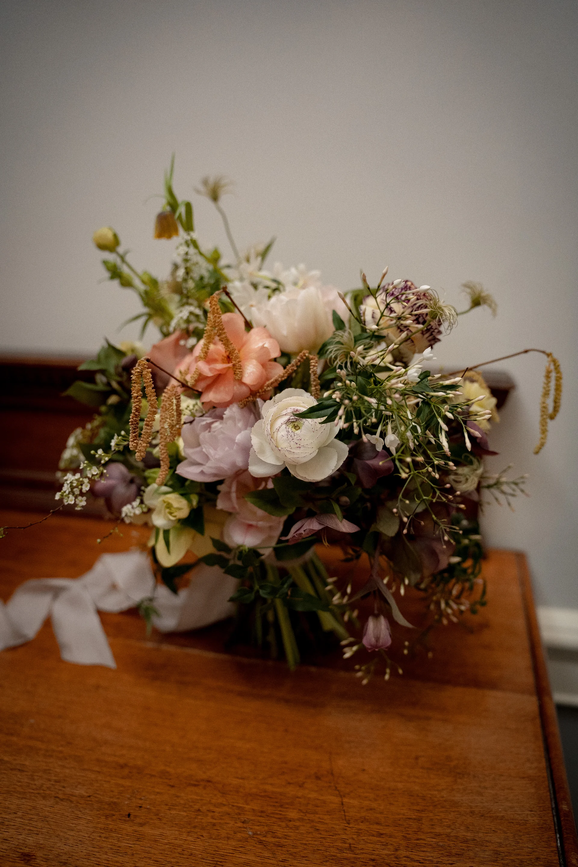 An unstructured garden-style bridal bouquet featuring pastel roses and ranunculus rests on a wooden surface with a trailing pale ribbon.