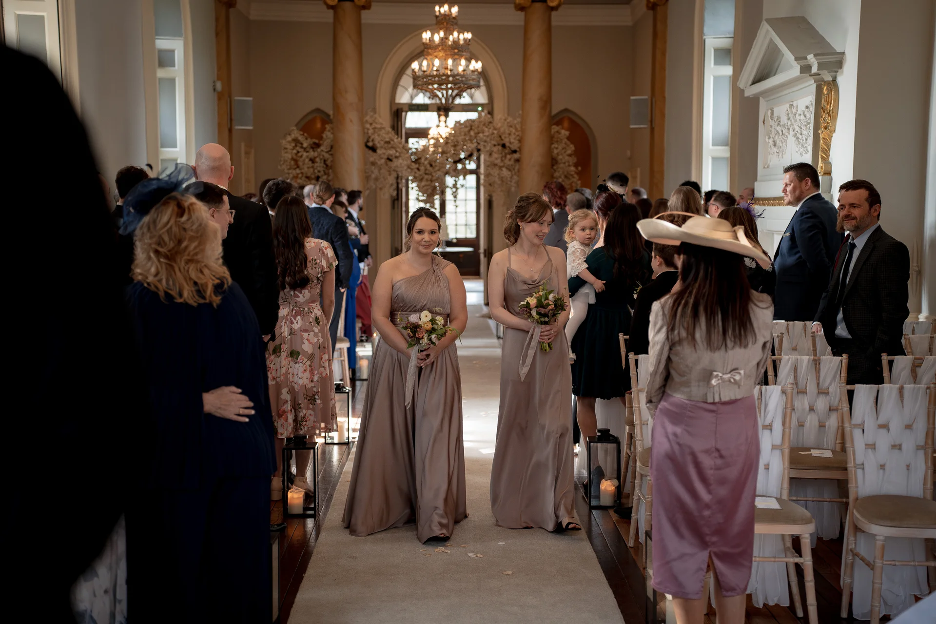 Two bridesmaids in matching satin taupe gowns walk down the aisle during a grand country house wedding ceremony.