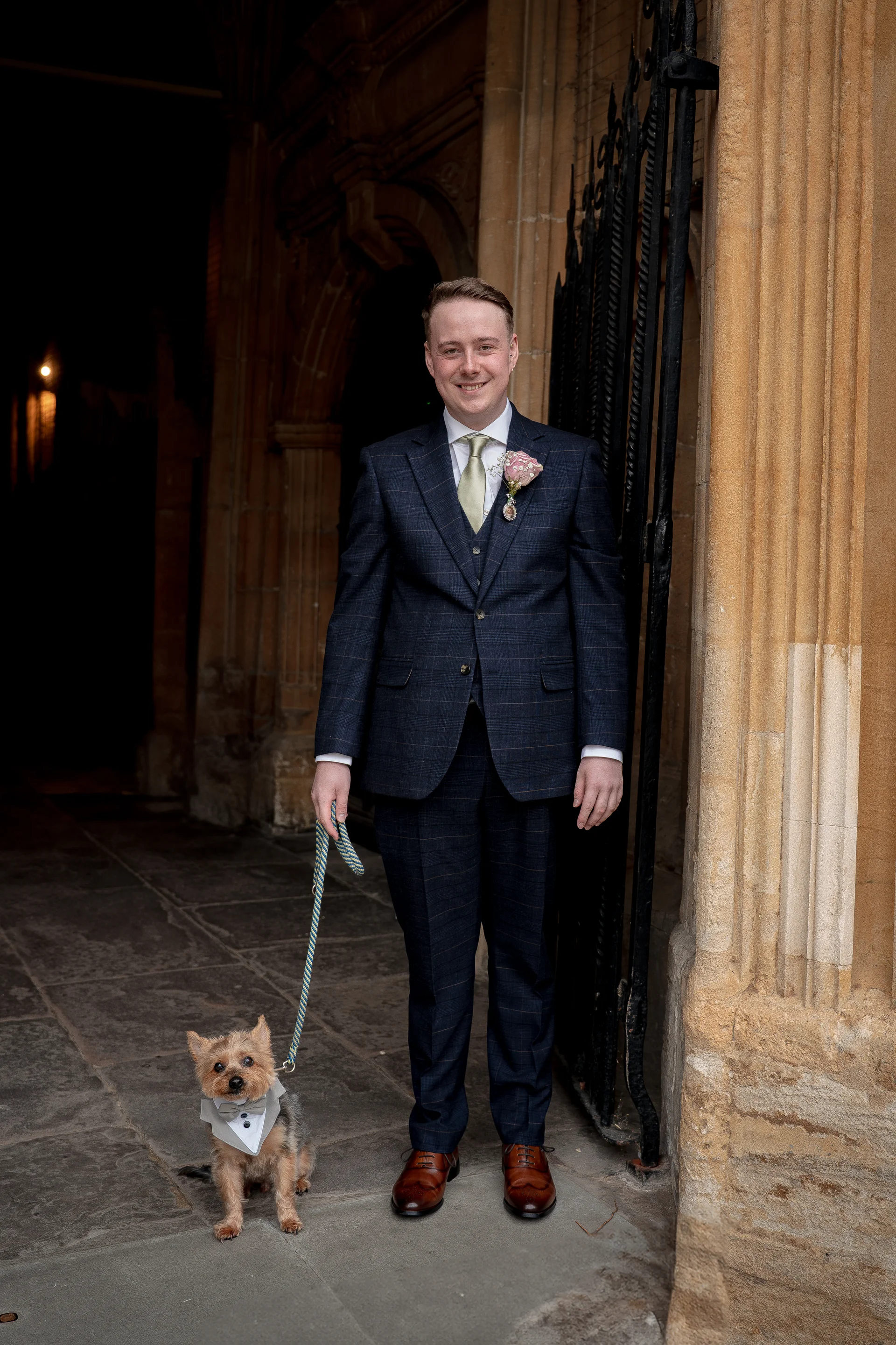 A smiling groom in a patterned blue suit stands next to a small dog wearing a mini tuxedo collar while holding its leash near an old stone church doorway.