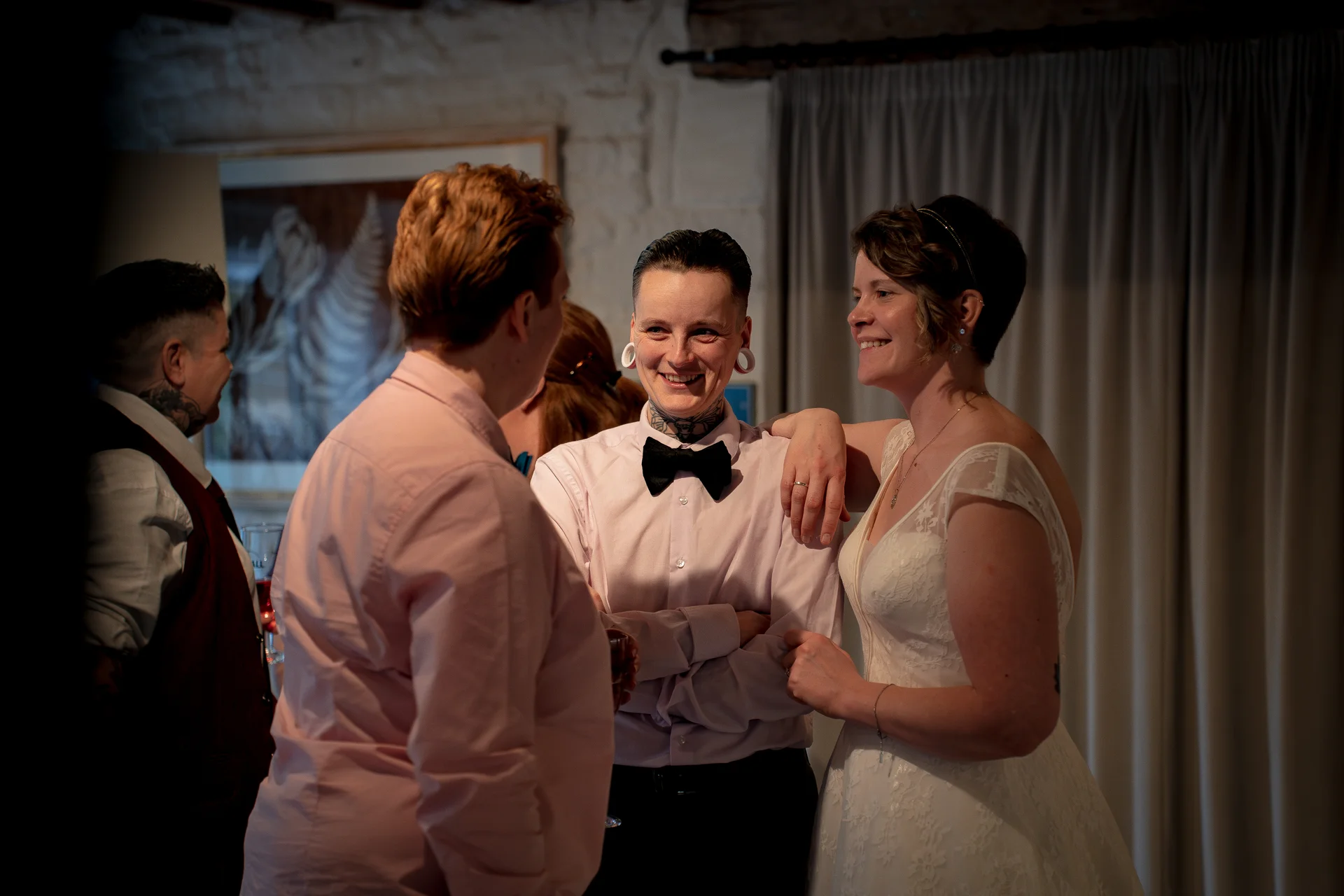 A smiling couple and wedding guests chat happily together indoors during a relaxed evening reception.