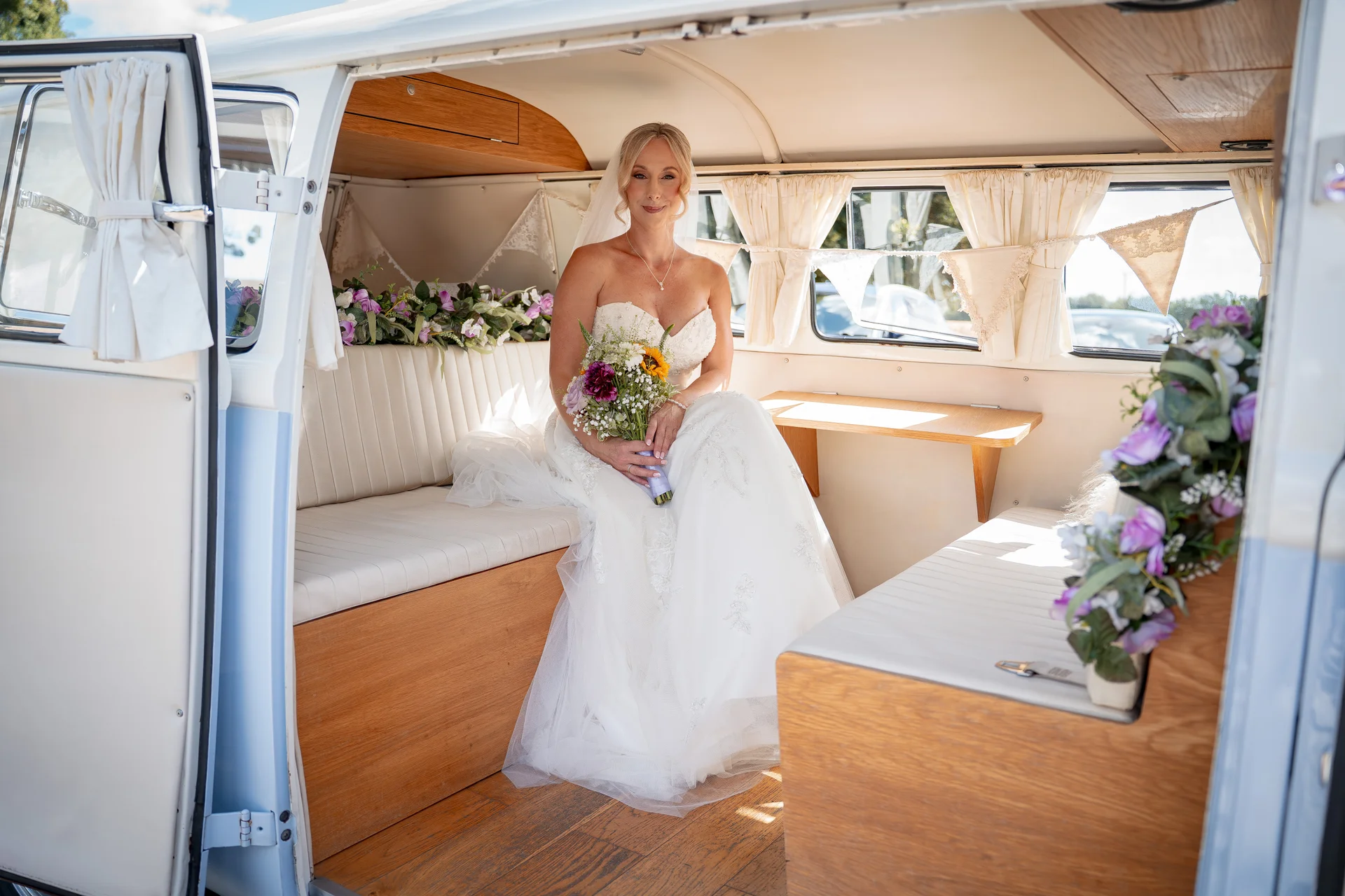A beautiful bride in a strapless white gown sits inside a vintage VW campervan, holding a colourful bouquet of flowers.