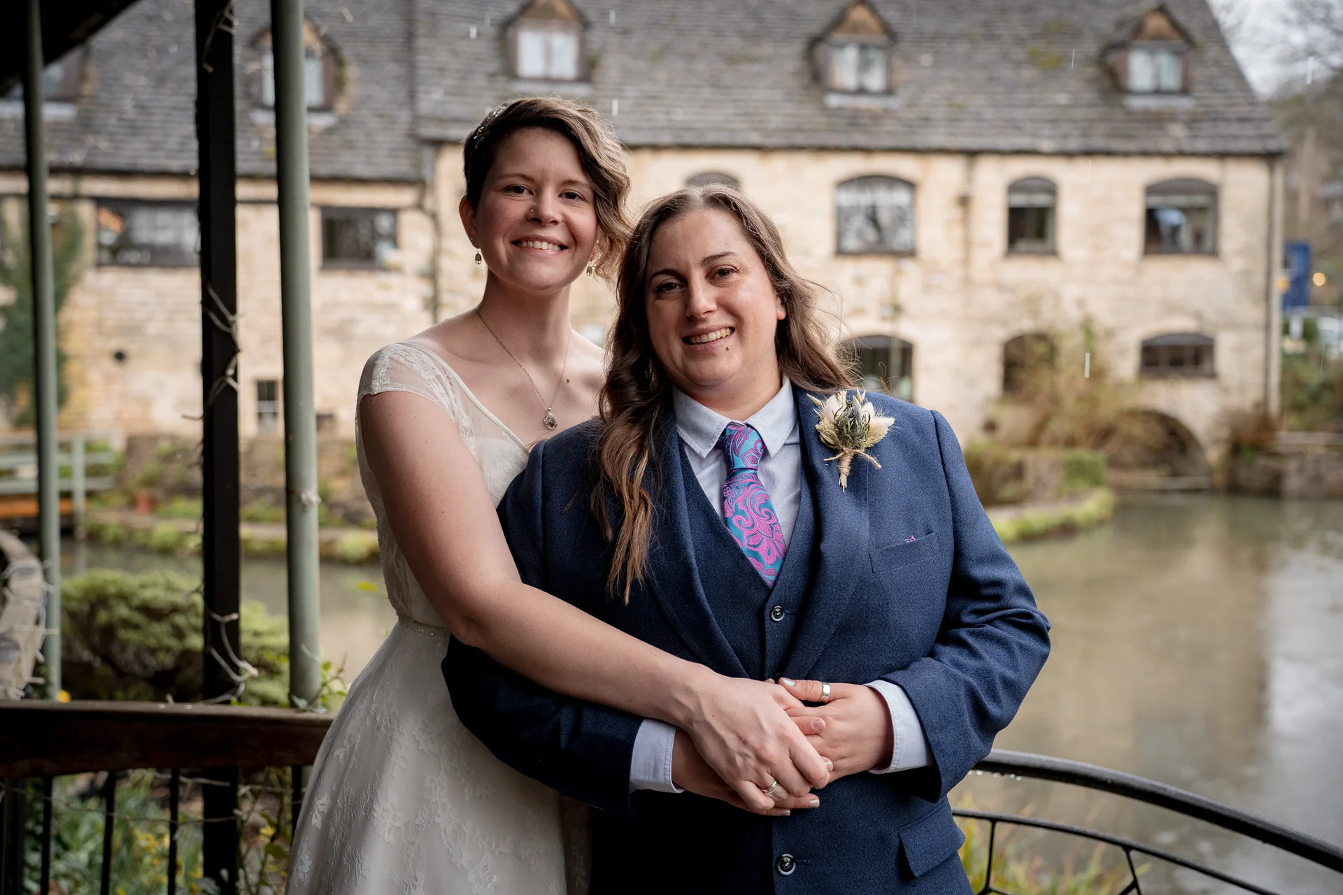 A happy same-sex couple poses together outdoors with one bride in a white lace dress and the other in a smart blue three-piece suit with a patterned tie and boutonniere, against the backdrop of a rustic stone building by the water.