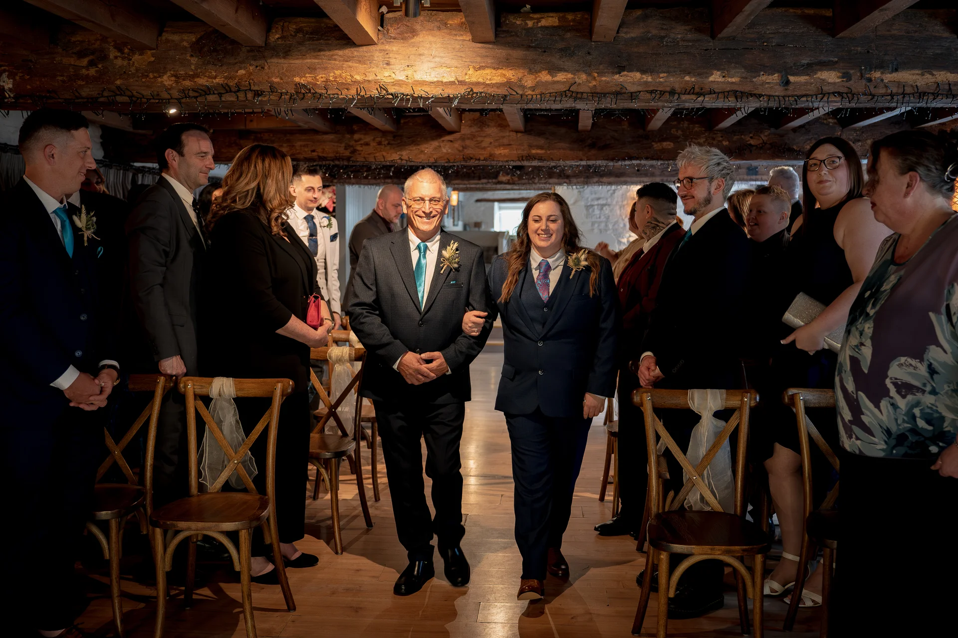 A smiling couple walks down the aisle arm in arm through a rustic barn venue decorated with exposed wooden beams and fairy lights, surrounded by cheering guests.