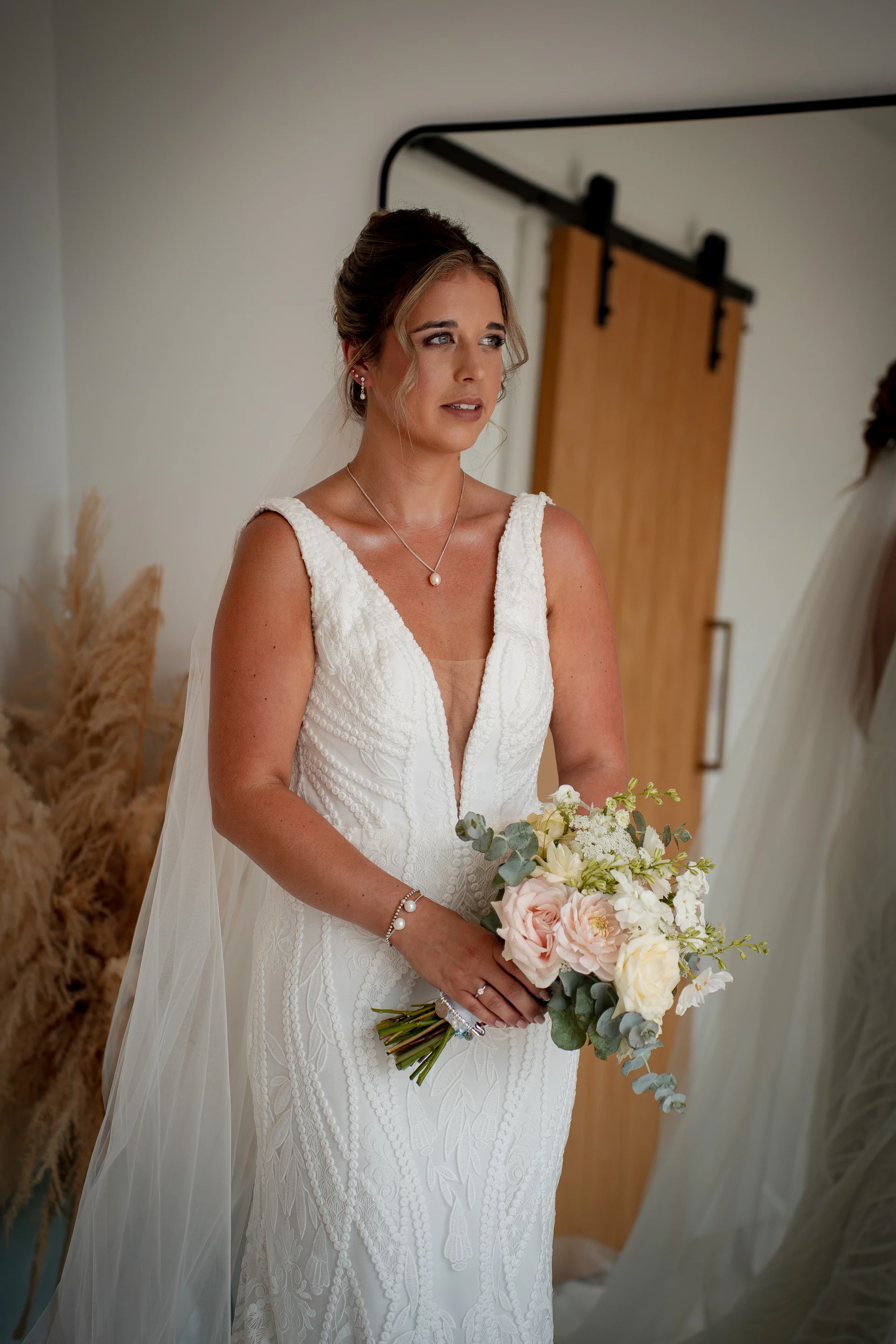 A glowing bride in a patterned V-neck lace wedding dress holds a delicate pastel bouquet while looking aside in a bright indoor room.