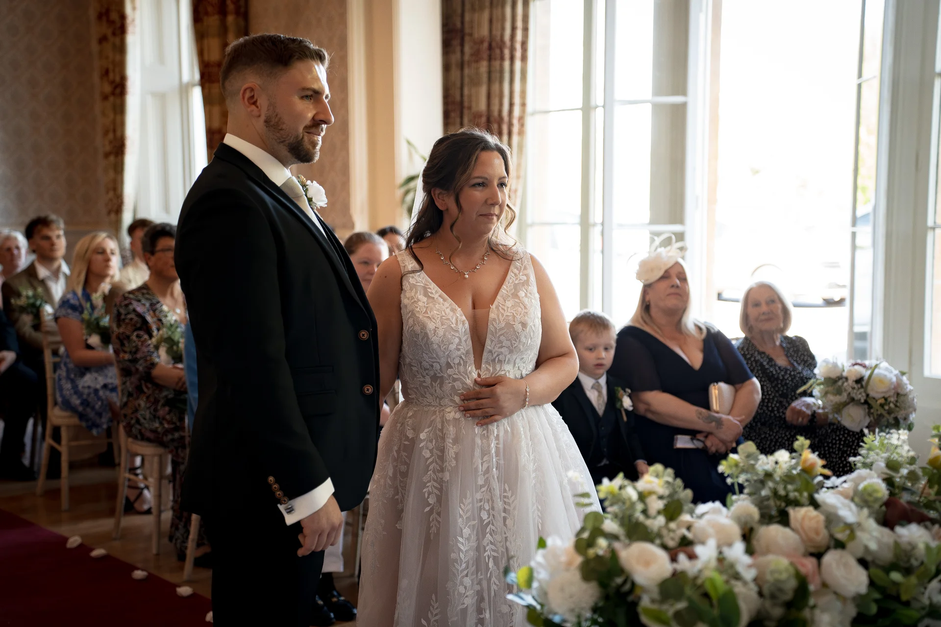A bride and groom stand together at the altar during their civil ceremony, surrounded by seated guests and elegant floral arrangements.