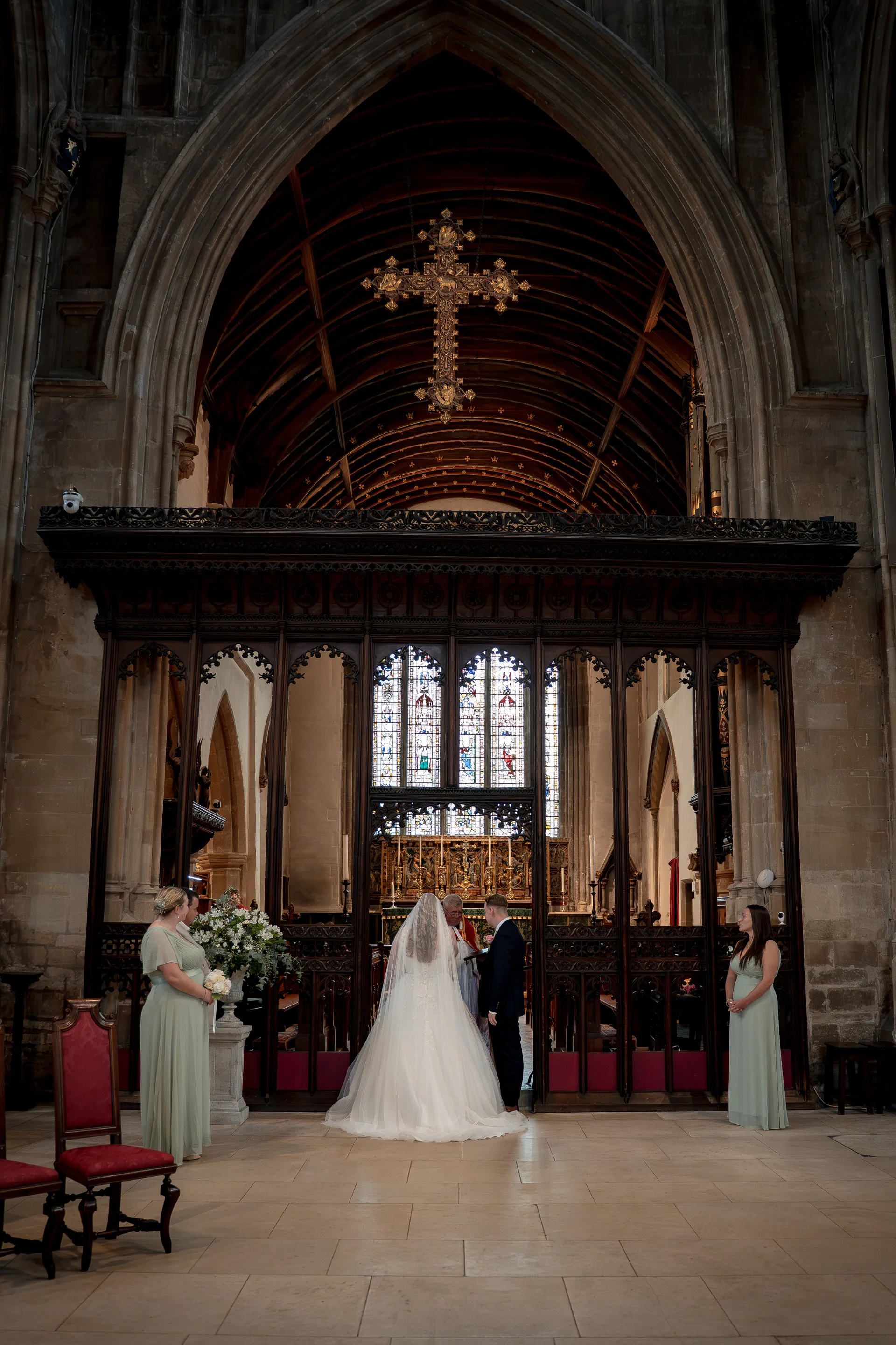 A wide angle shot of a traditional church wedding ceremony featuring the bride in a classic white gown with a long veil standing at the altar with the groom, flanked by bridesmaids in sage green dresses beneath a magnificent wooden rood screen and stone archway.