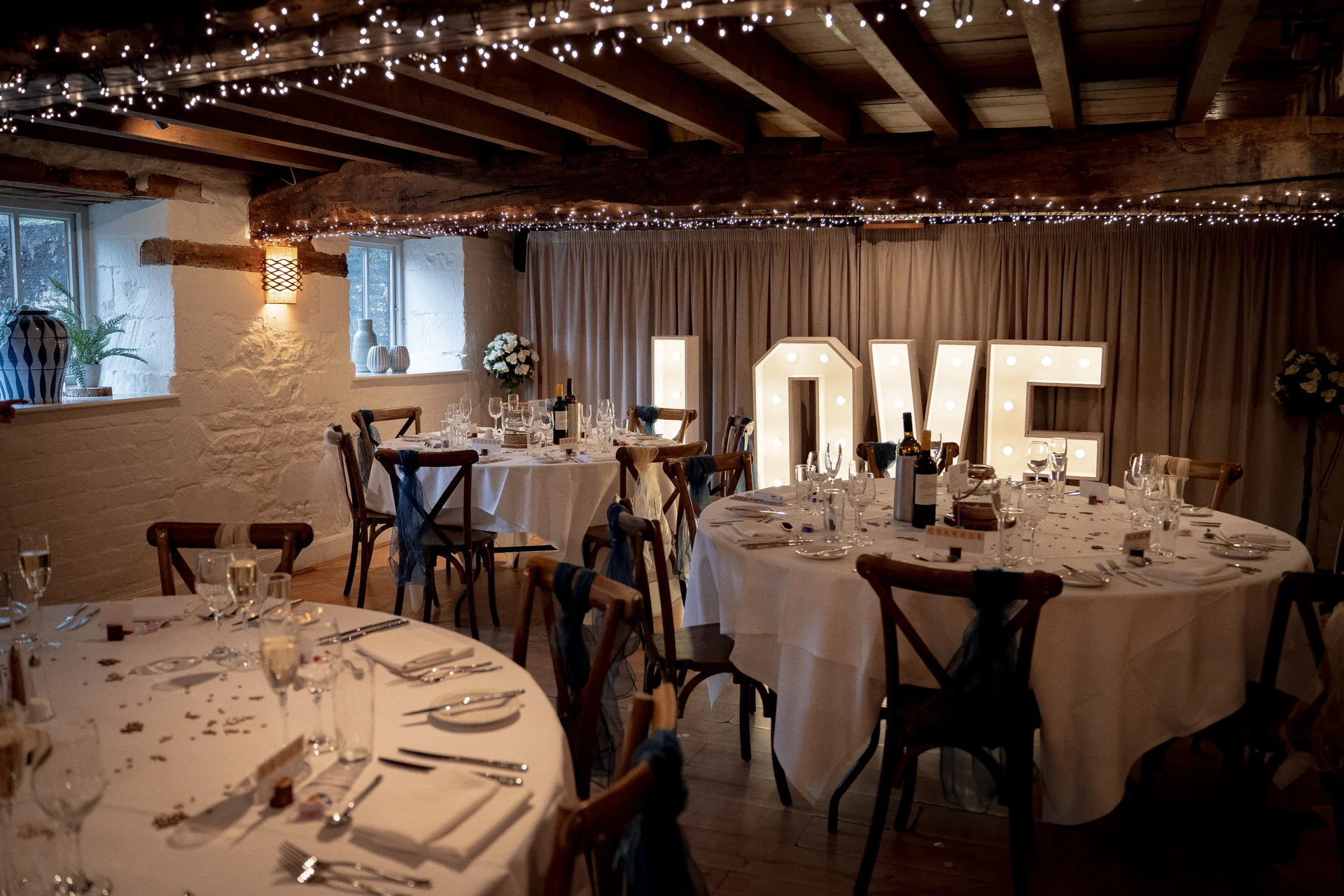 A rustic barn wedding reception featuring round guest tables with crisp white linens and glowing fairy lights strung across exposed wooden ceiling beams.
