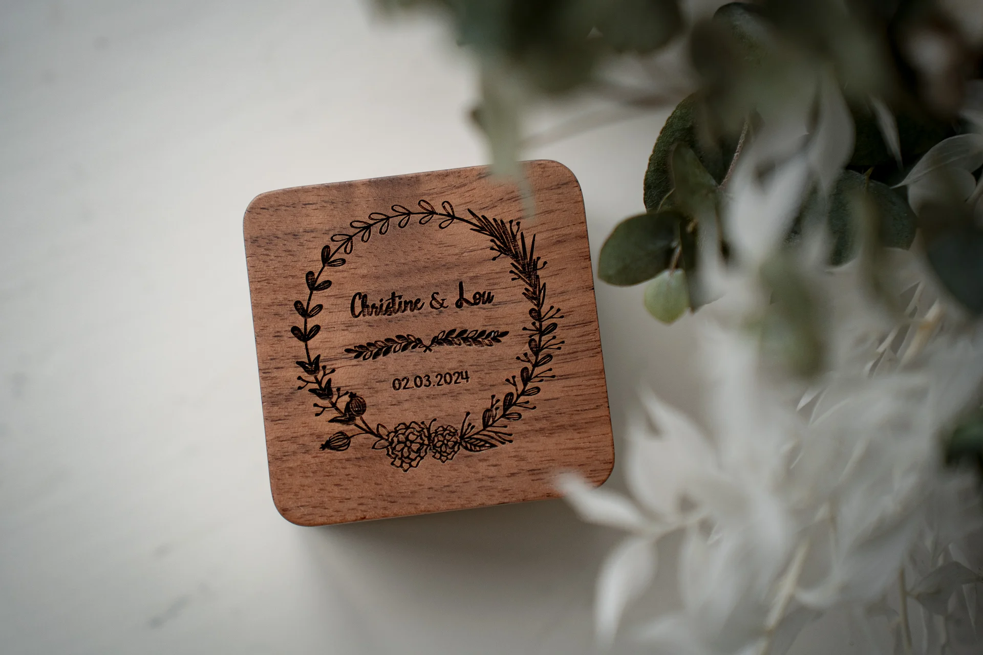 A close up detail shot of a personalised wooden ring box featuring engraved botanical wreaths and a wedding date, resting beside delicate dried foliage.