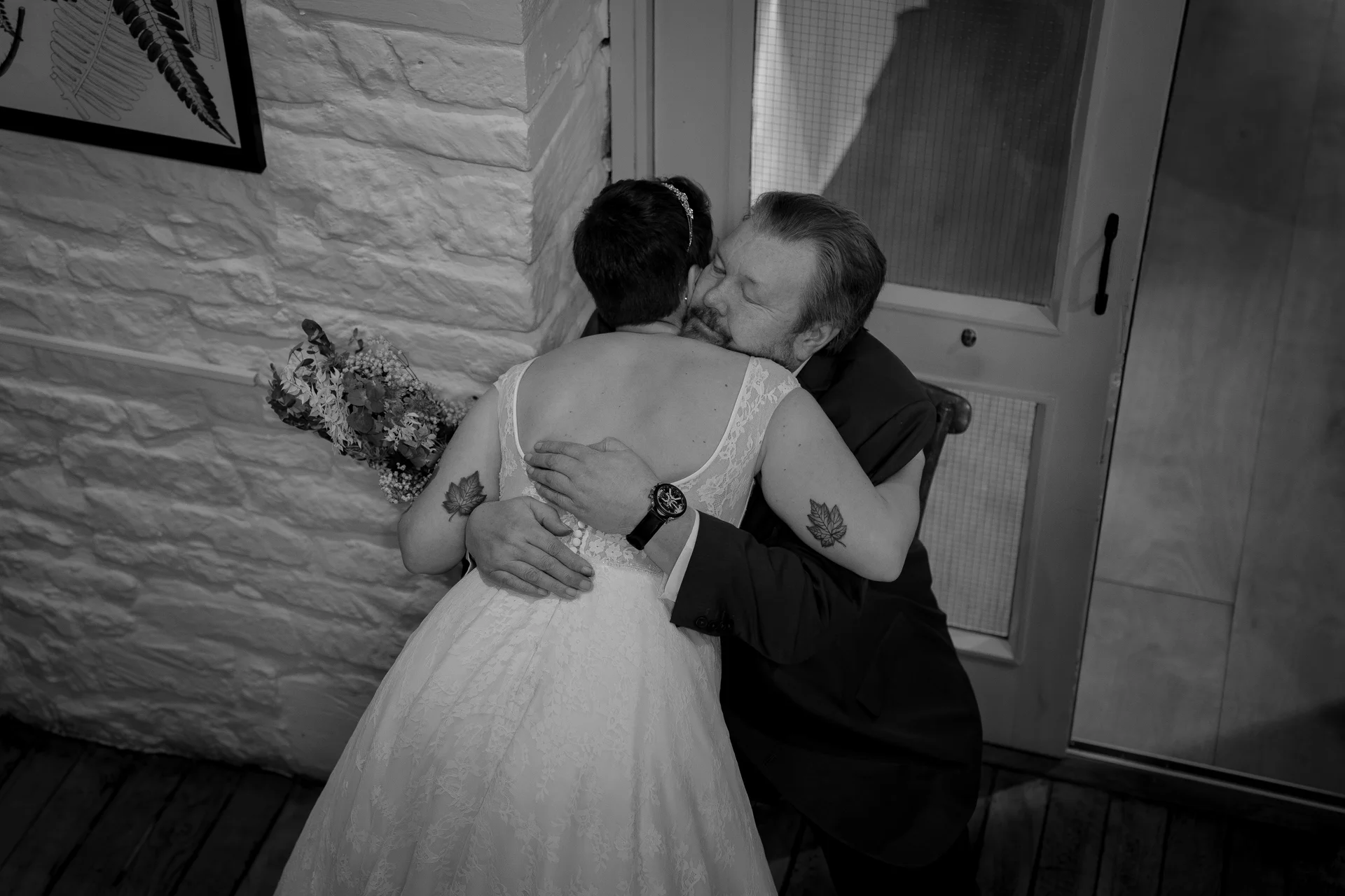 A tender black and white candid shot of a short-haired bride in a lace wedding dress sharing an emotional embrace and first look with her father indoors.