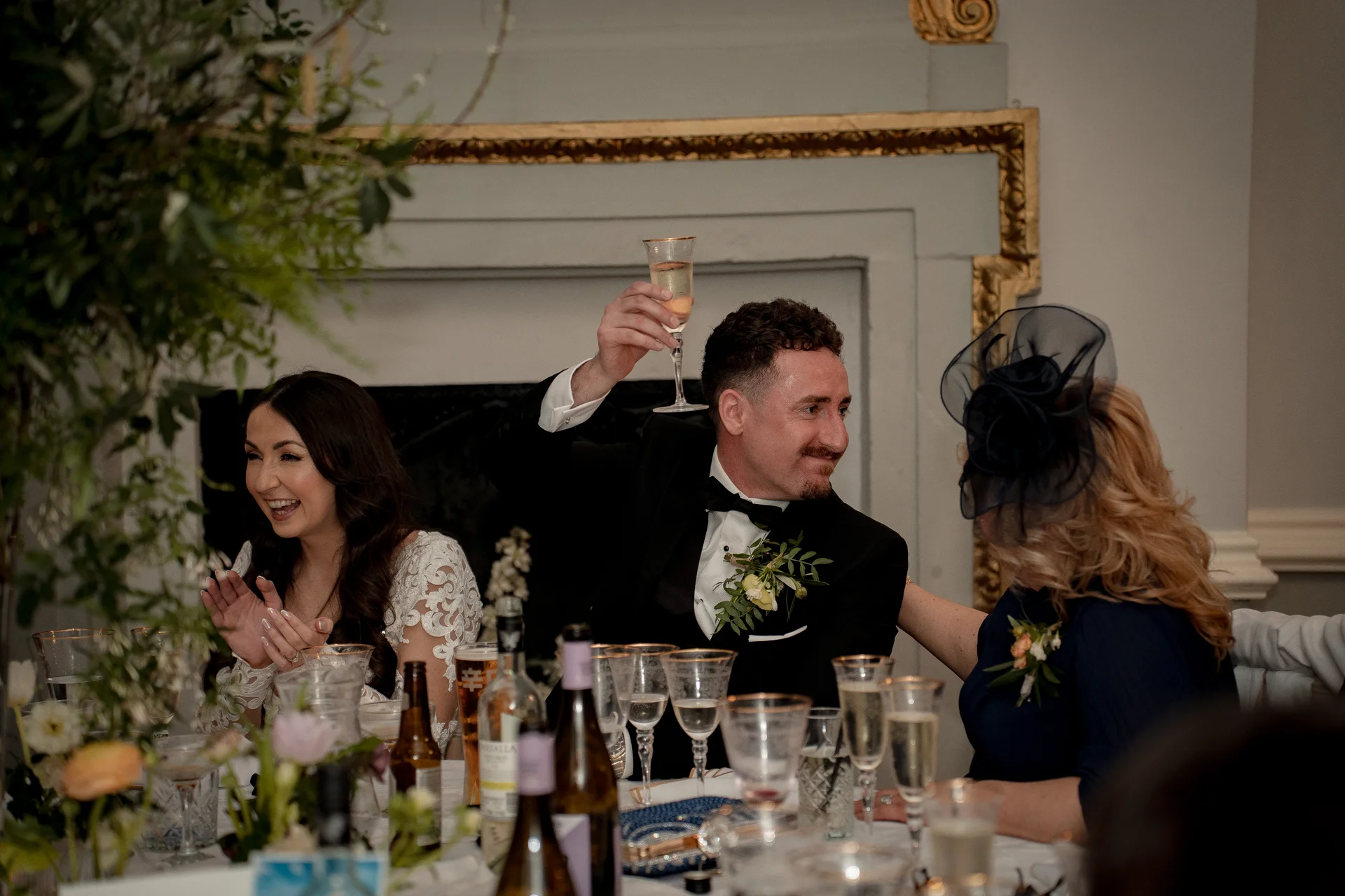 A groom in a black tuxedo raises a champagne flute to toast during wedding speeches, while the bride and a guest laugh and applaud at the top table.