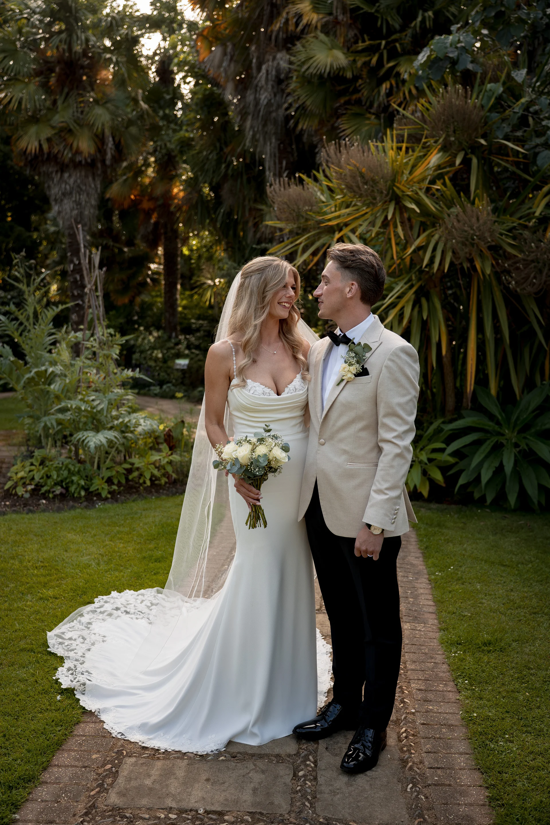 A smiling bride in a cowl neck crepe gown with a long train and veil looks lovingly at her groom in a cream jacket standing on a garden pathway.