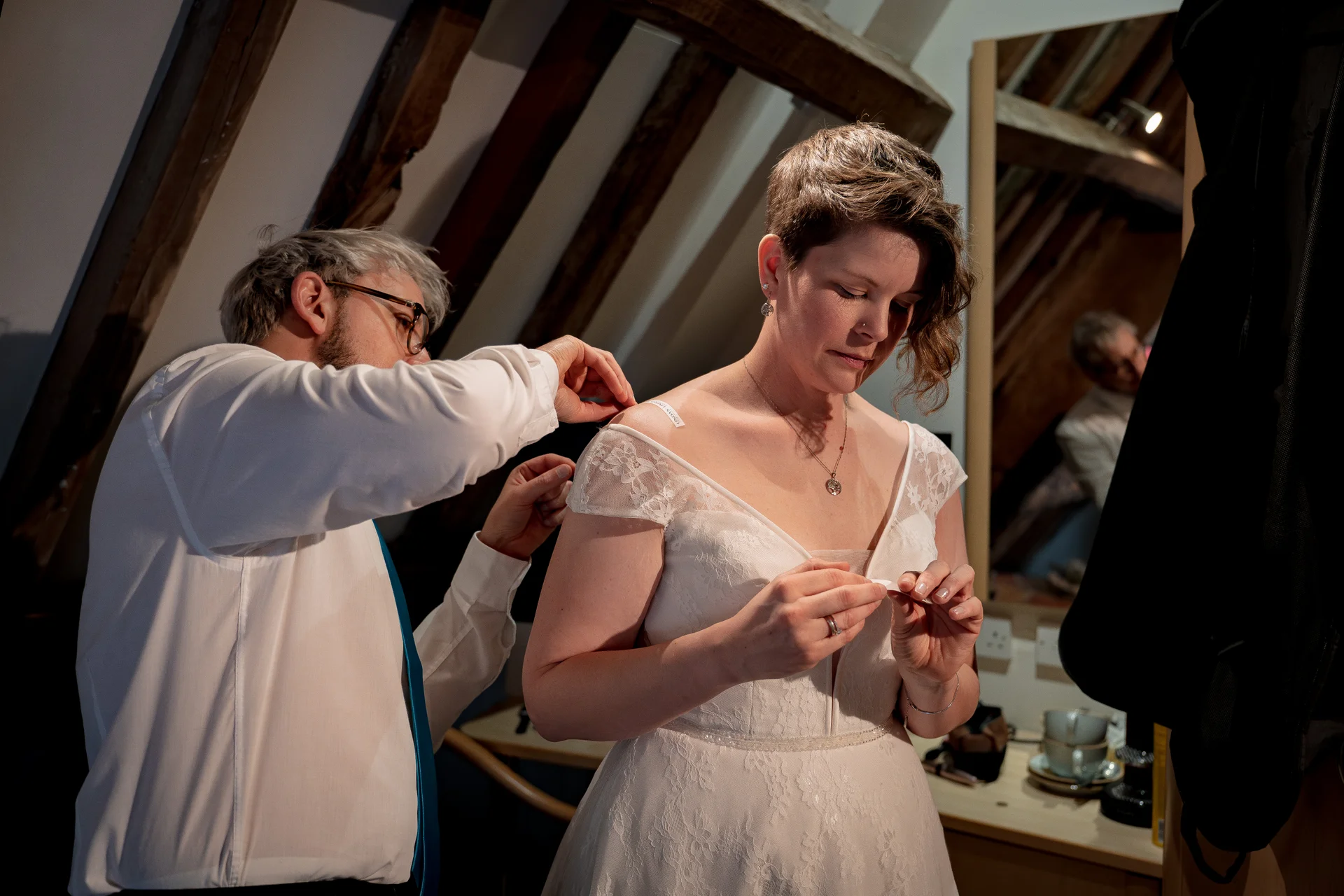 A bride with short hair adjusts her lace wedding dress while getting ready indoors under exposed wooden rafters.