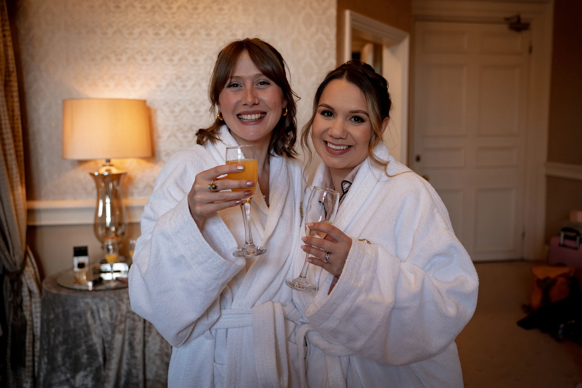 Two smiling brides in white bathrobes hold champagne flutes and toast each other during morning wedding preparations in a hotel room.