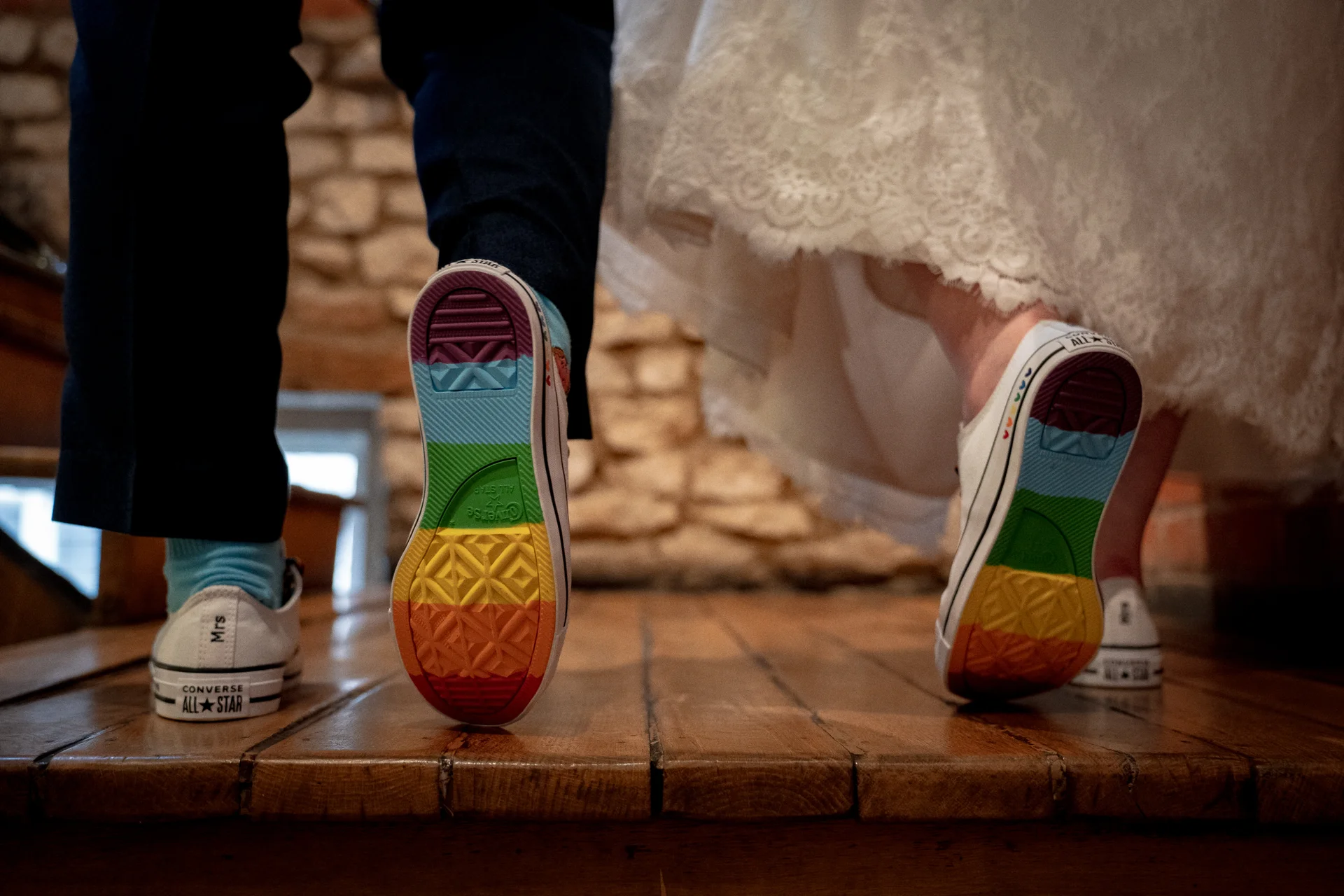 A low-angle shot of a newlywed couple showing off their matching white Converse trainers with colourful rainbow soles while walking across a wooden floor.