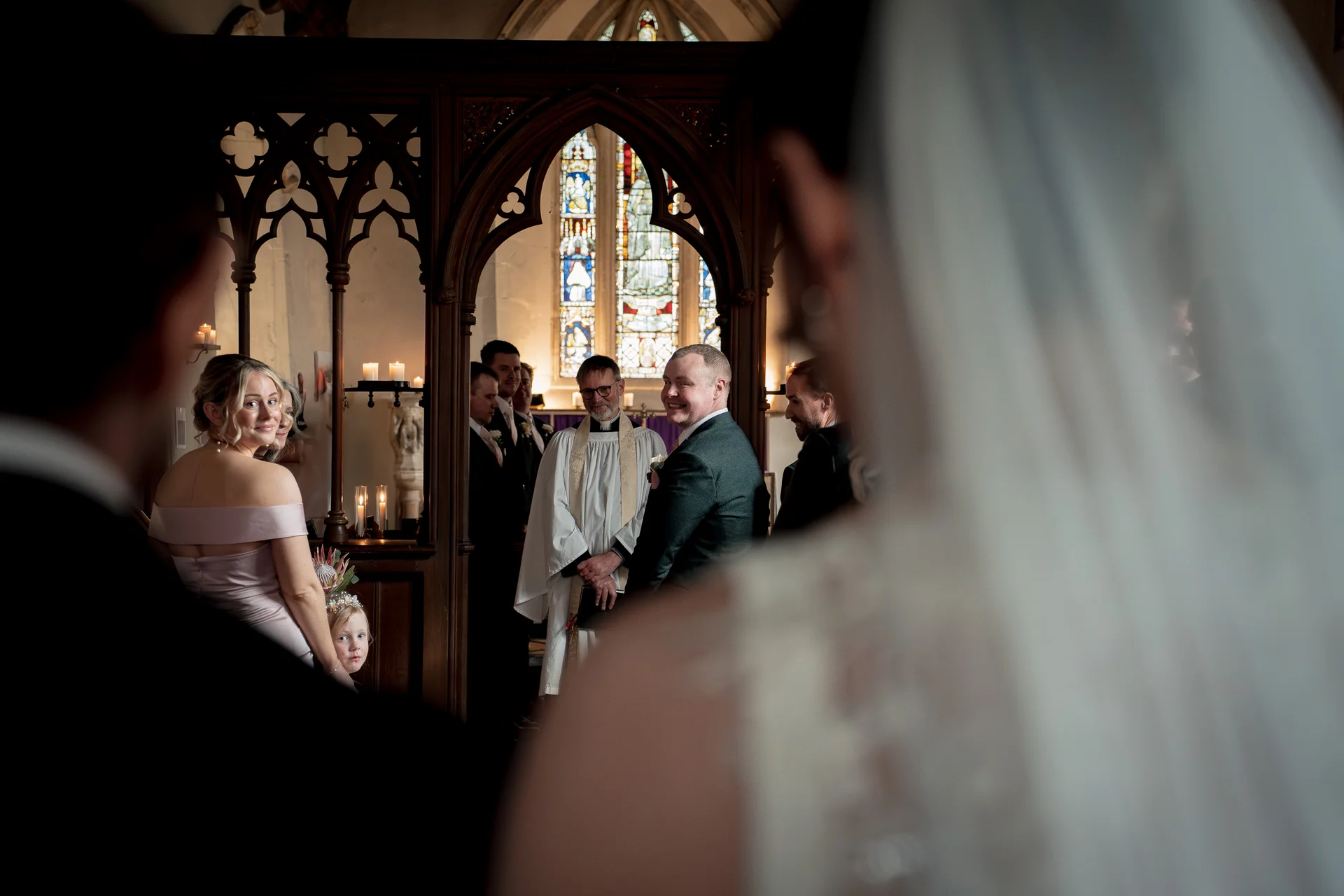 An over the shoulder perspective looking through a wooden screen towards a groom and officiant standing near stained glass windows inside a historic church.