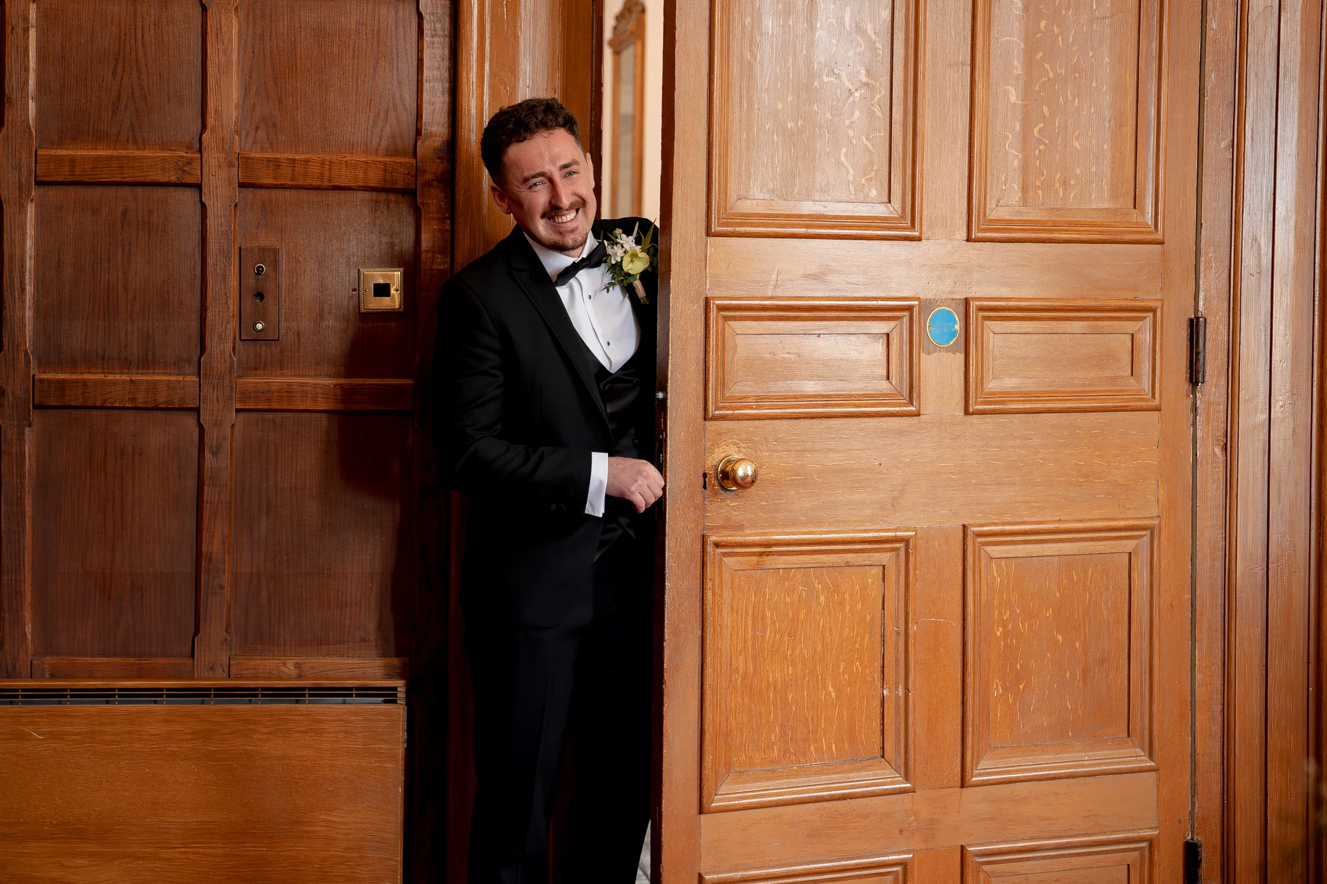 A smiling groom wearing a black tuxedo and bow tie peeks playfully through a heavy wooden panelled door.