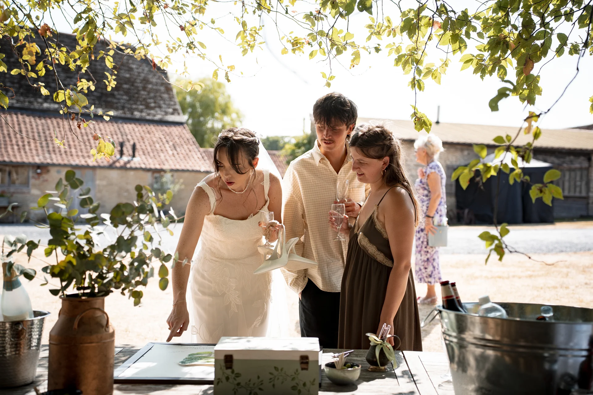 A bride in a white gown and guests gather around an outdoor table in a sunny barn courtyard, holding drinks and enjoying the wedding celebration.