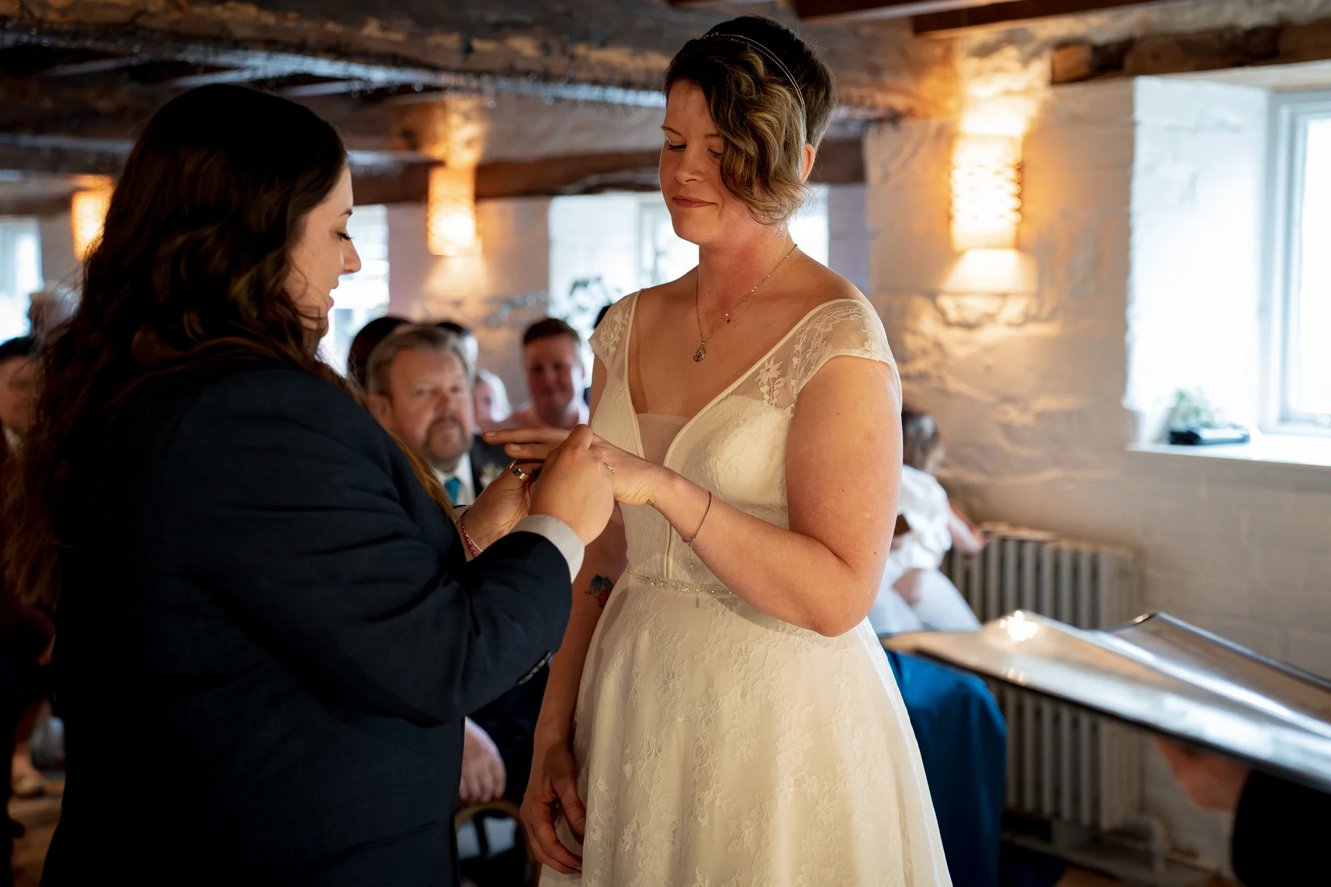 Two brides exchange rings during their civil ceremony inside an intimate stone barn venue illuminated by warm wall lighting.