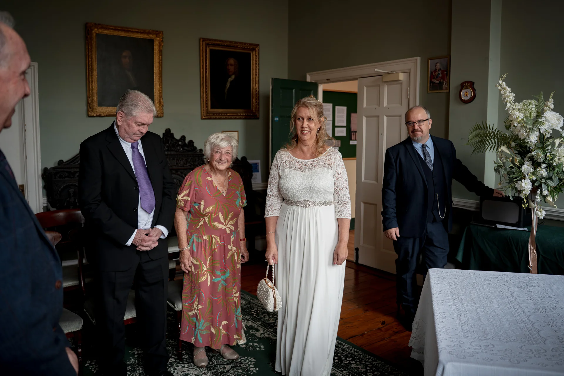 A mature bride in a lace-detailed a-line gown stands alongside family members inside a traditional historic venue featuring framed portraits.
