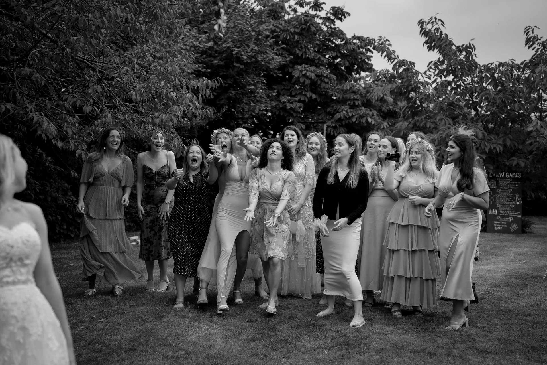 A black and white photograph captures a bride throwing her bouquet to an enthusiastic group of bridesmaids and guests gathered on a lush garden lawn.