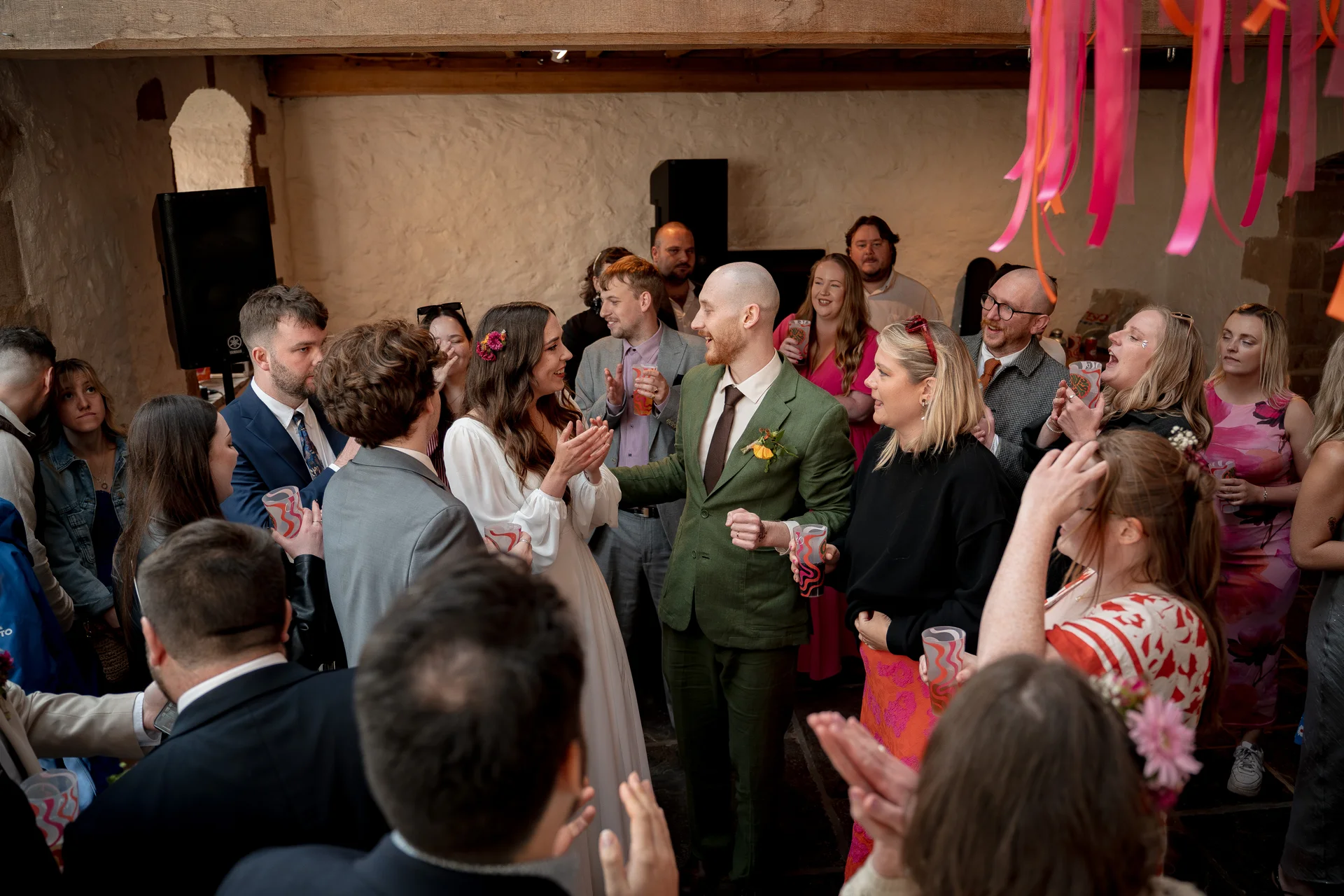 Newlyweds in a rustic stone barn are joyfully applauded by their wedding guests holding drinks beneath hanging pink ribbons.