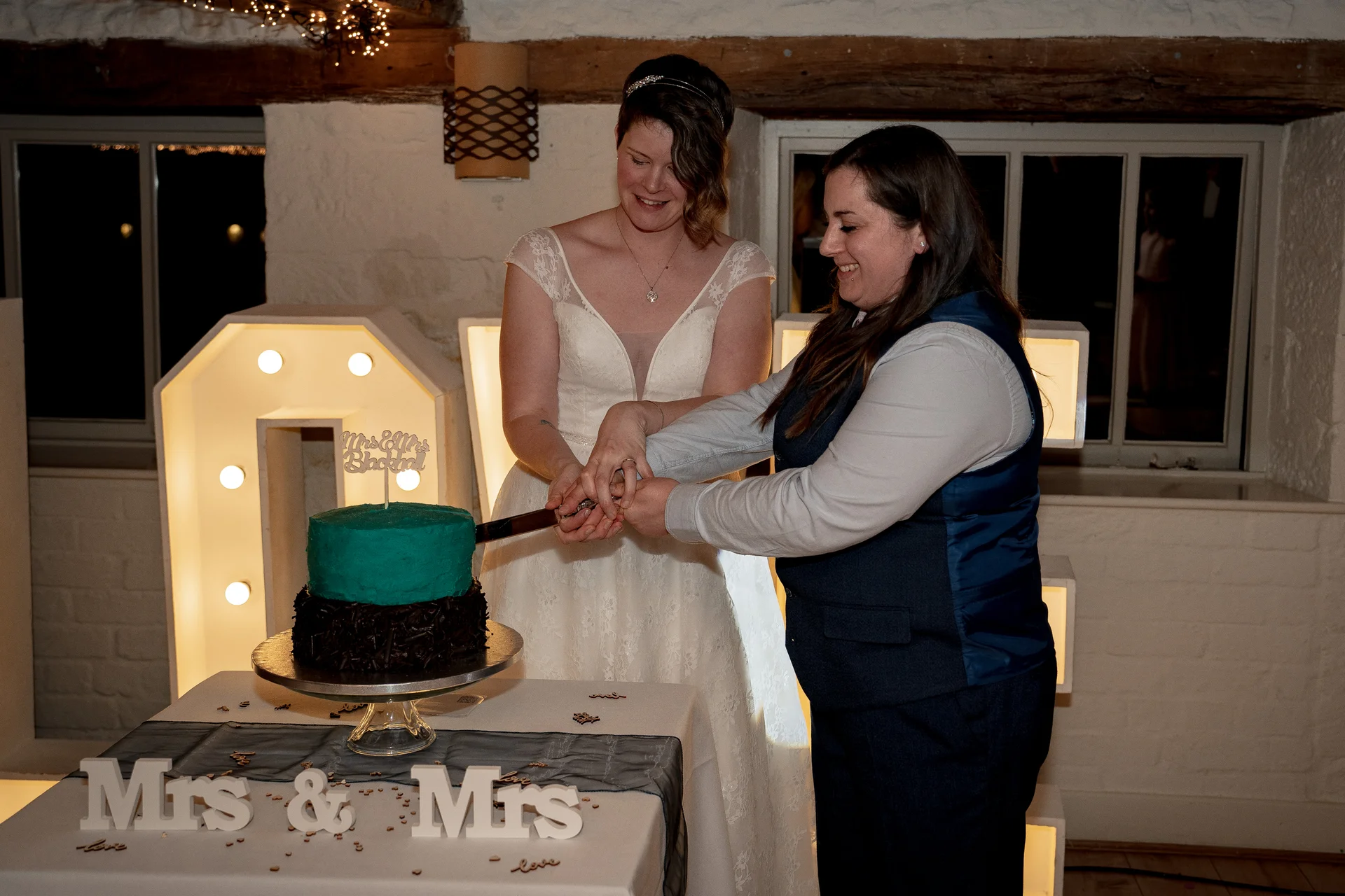 Two brides smiling together as they cut a two-tier teal wedding cake in a rustic barn venue illuminated by large light-up letters.