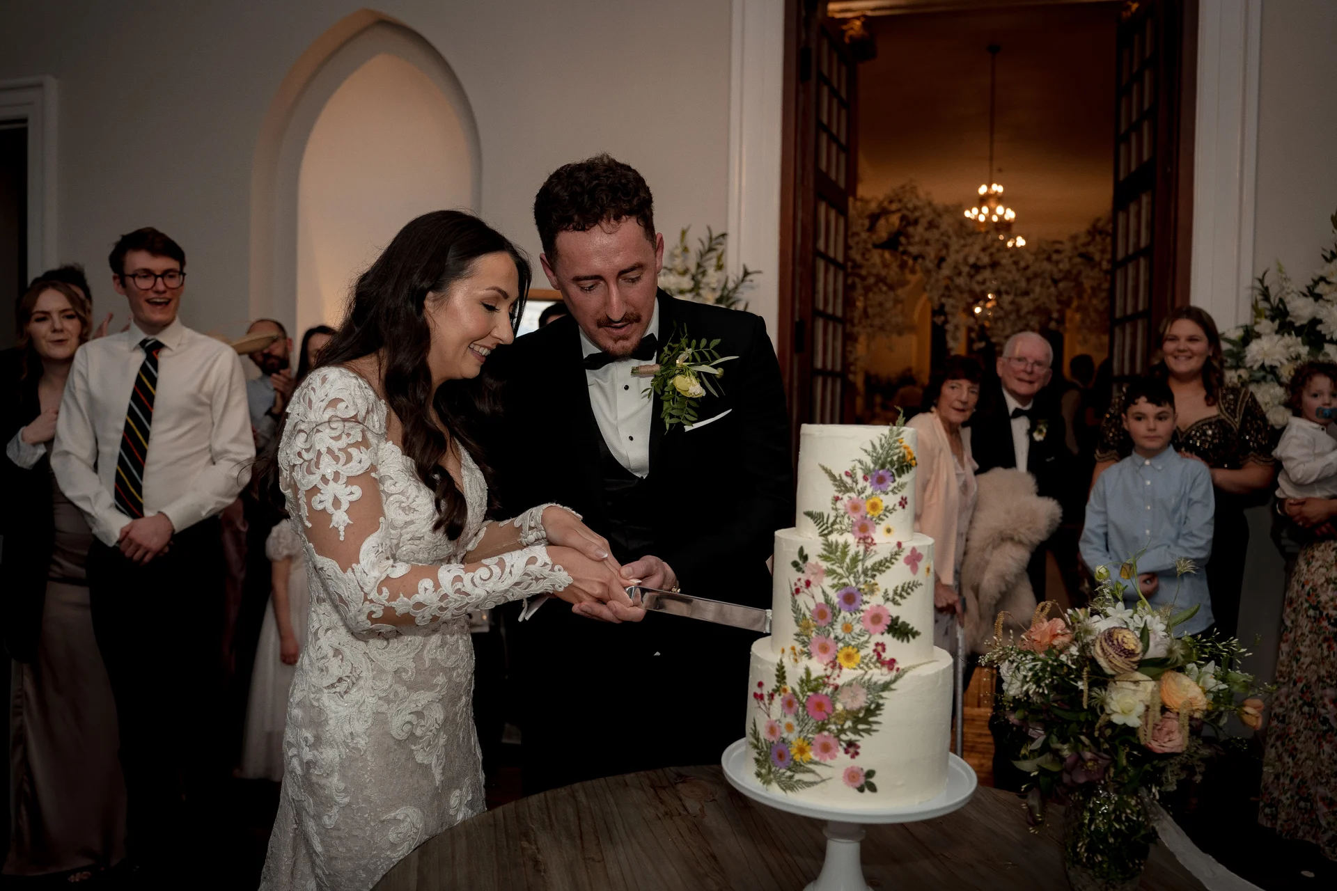 The bride and groom smile as they cut a three-tiered white wedding cake decorated with pressed colorful wildflowers, surrounded by cheering guests.