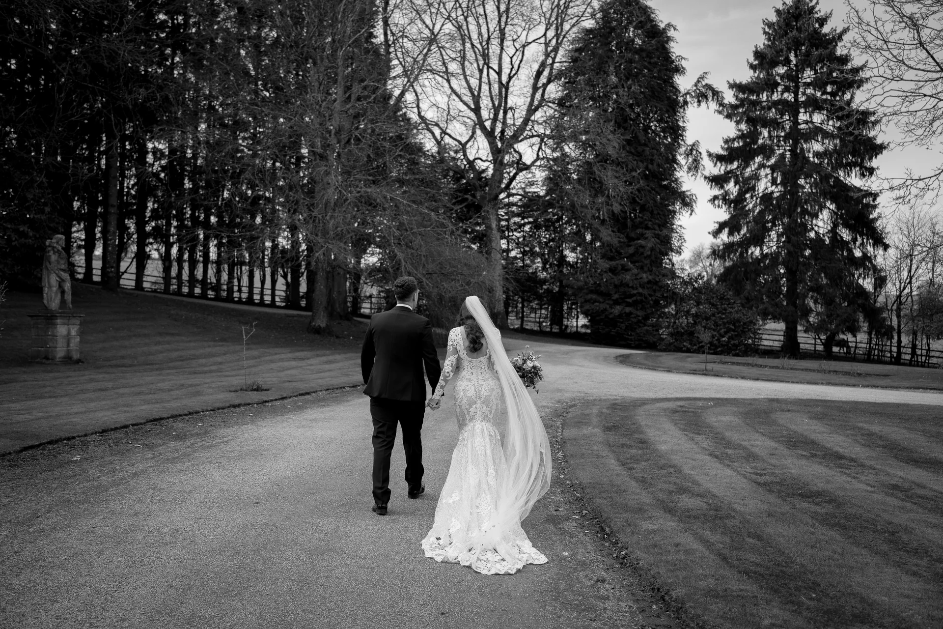 Newlywed bride and groom walk hand in hand down a sweeping estate driveway lined with tall trees, captured from behind in a dramatic black and white photograph.