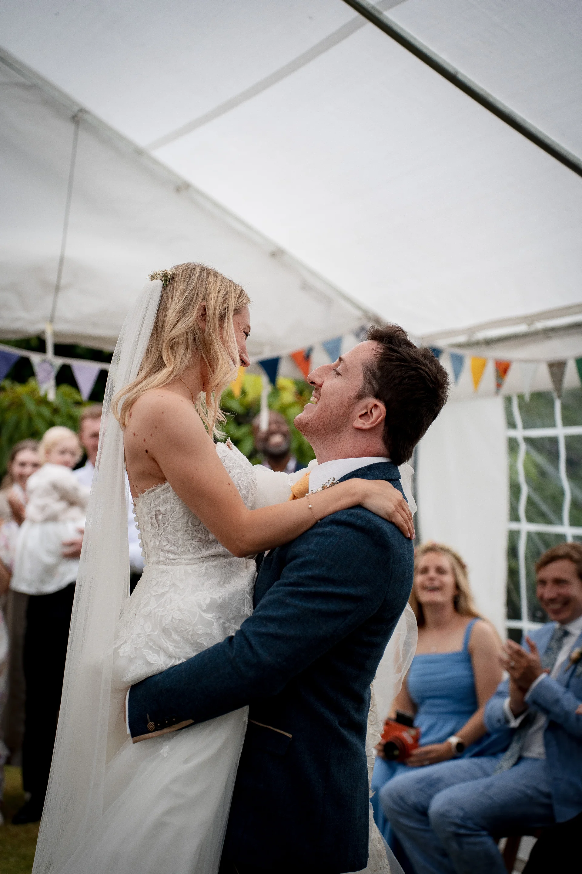 A smiling groom lifts his laughing bride into the air inside a marquee decorated with bunting, while guests cheer in the background.