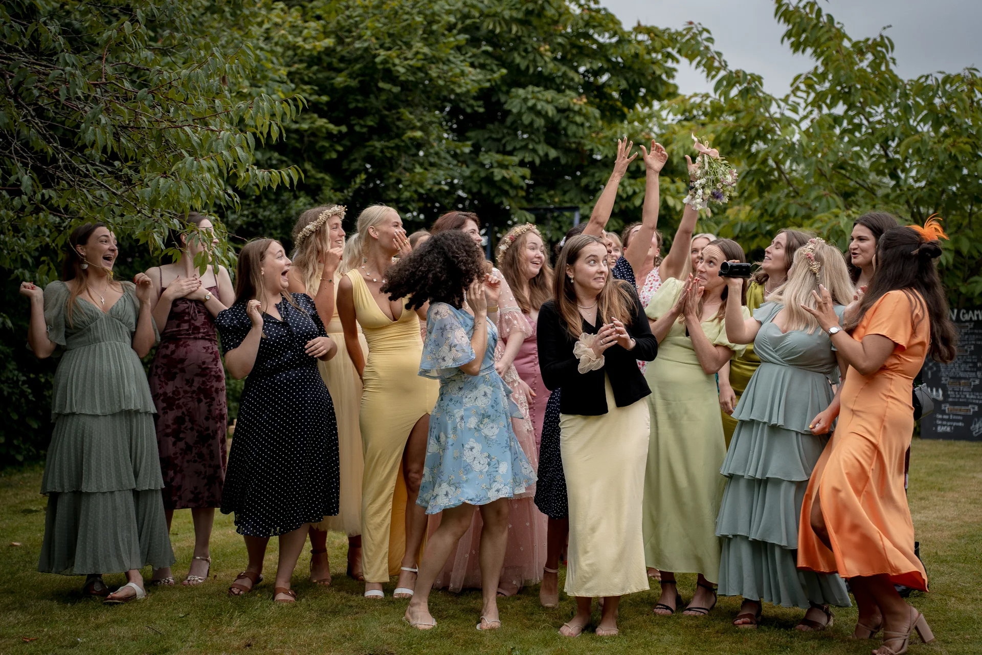 A lively group of bridesmaids and wedding guests react with joy and excitement as a bouquet is tossed into the air outdoors beneath lush green trees.