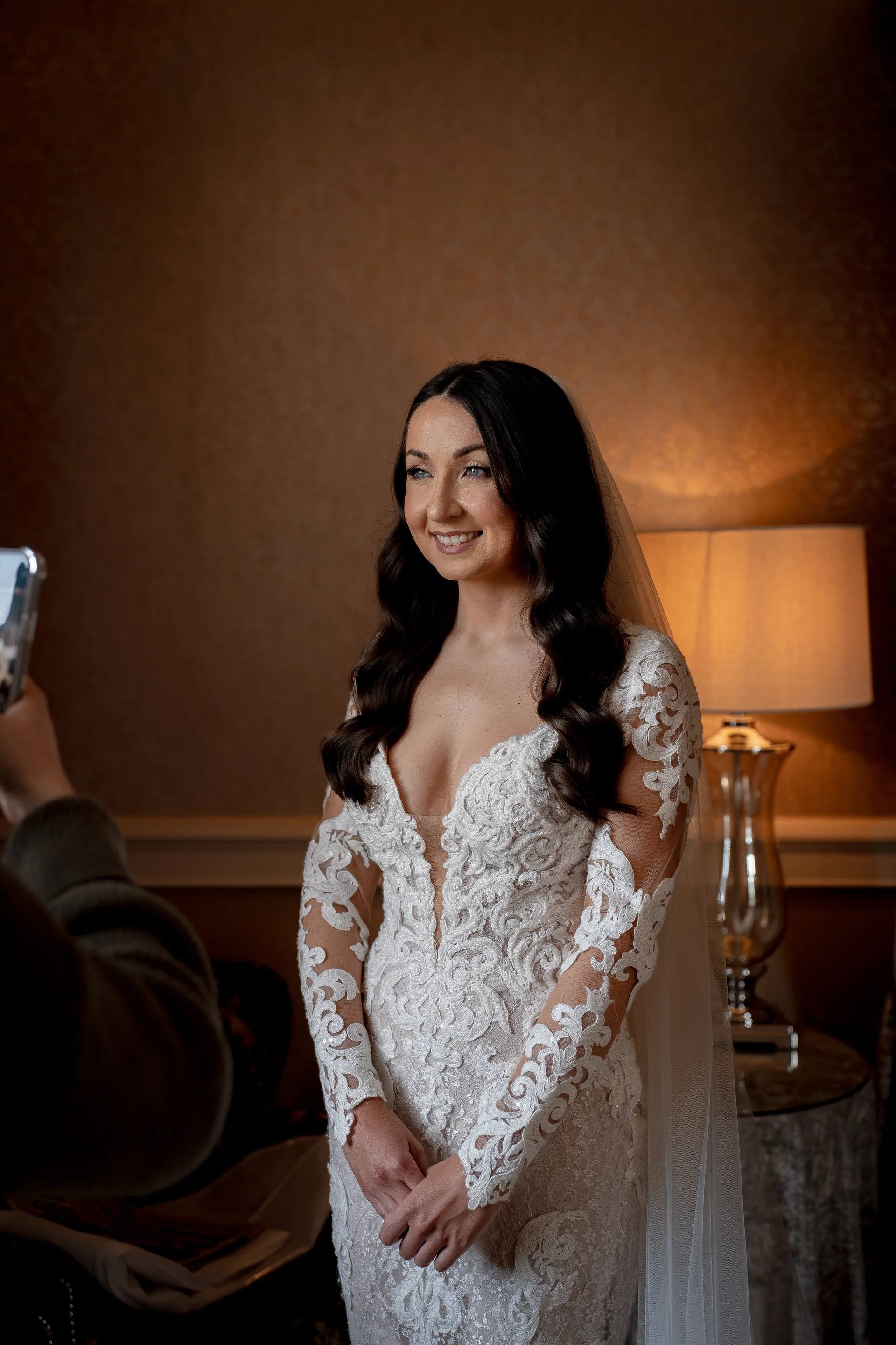 A smiling bride wearing a sheer lace long-sleeve gown and veil stands indoors while someone captures her portrait on a smartphone.