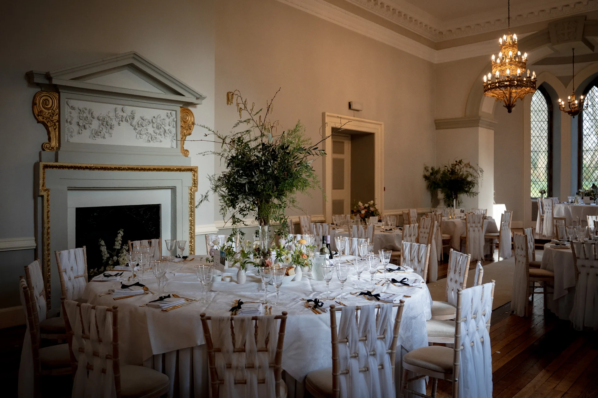 An elegant country house wedding reception featuring beautifully styled round tables with tall greenery centrepieces, white linens, and grand chandeliers hanging from a high ceiling.