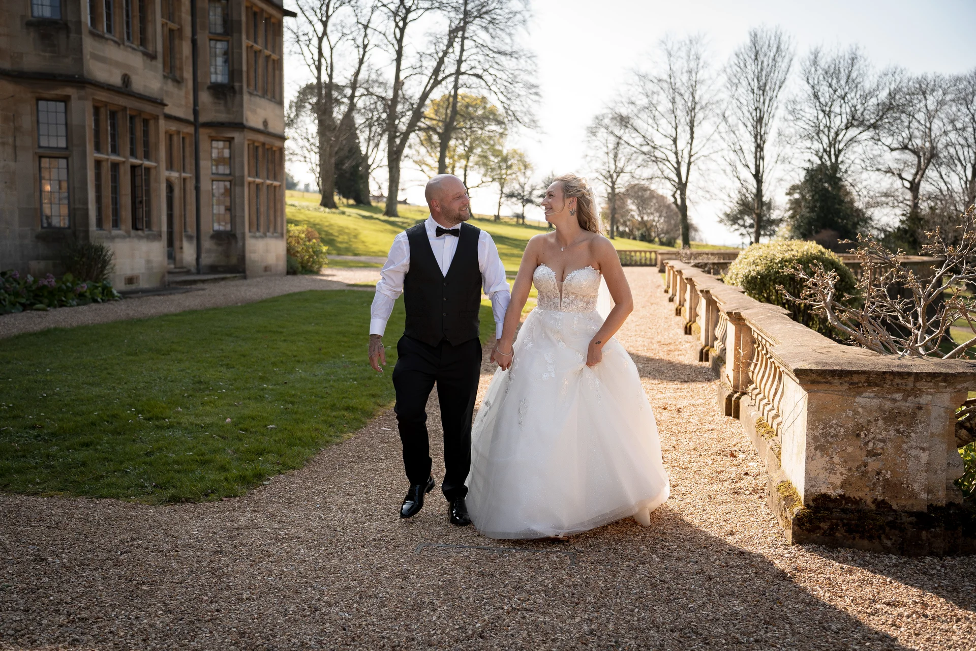 Newlyweds smiling at each other while walking hand in hand along a gravel path next to a historic stone manor house during golden hour.