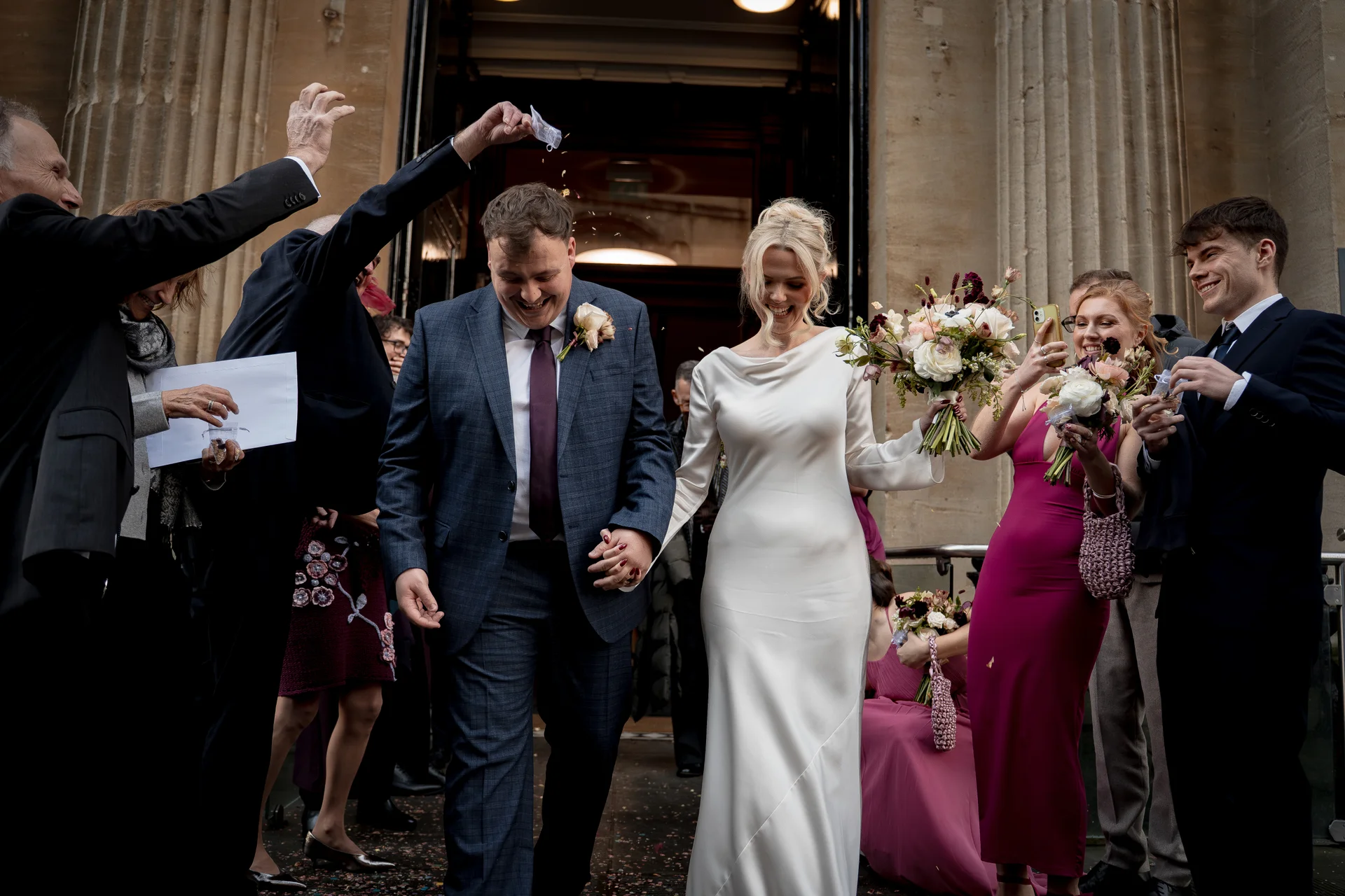 Newlyweds joyfully walk out of a stone building hand in hand as guests shower them with confetti during their wedding exit.