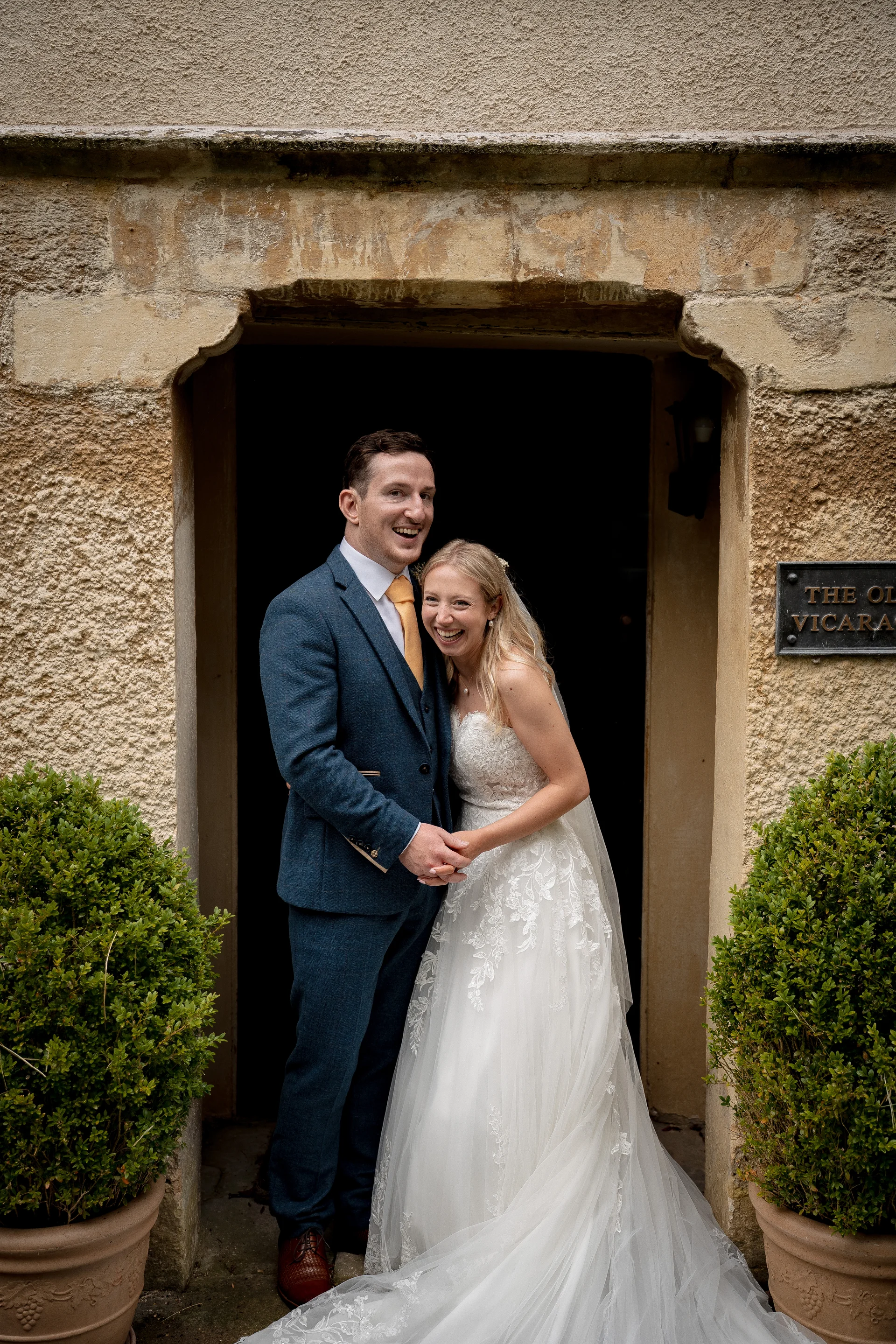 A joyful newlywed couple stand smiling hand in hand inside a stone doorway framed by potted topiary shrubs.