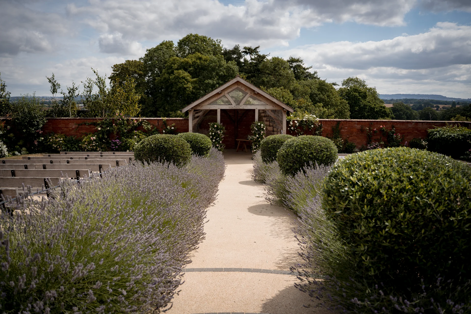 An outdoor wedding ceremony setup features a gravel aisle lined with rows of blooming lavender and topiary bushes leading towards a wooden gazebo framed by a brick wall.