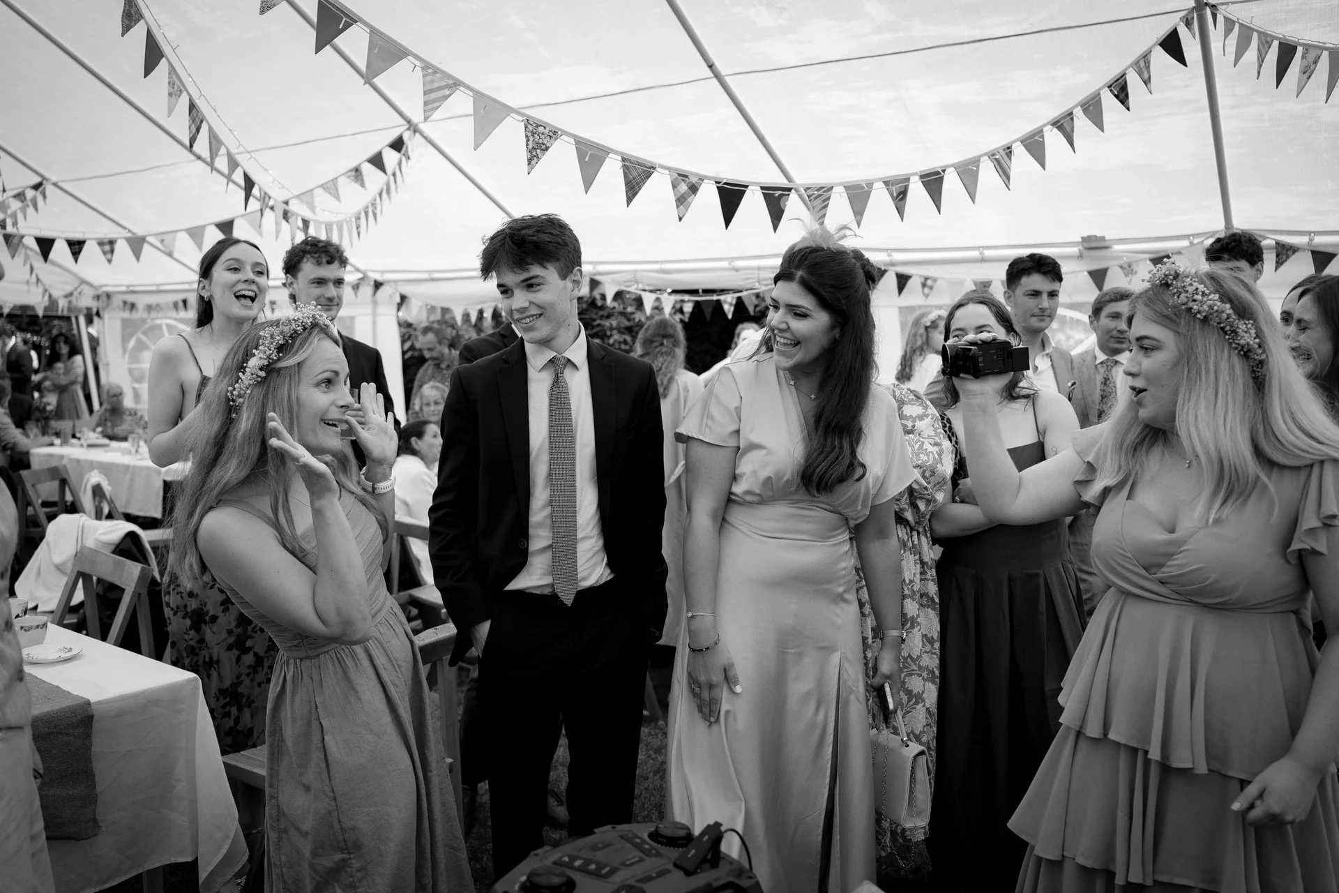 A joyful group of wedding guests wearing smart attire and floral crowns laugh together under a marquee decorated with hanging bunting.