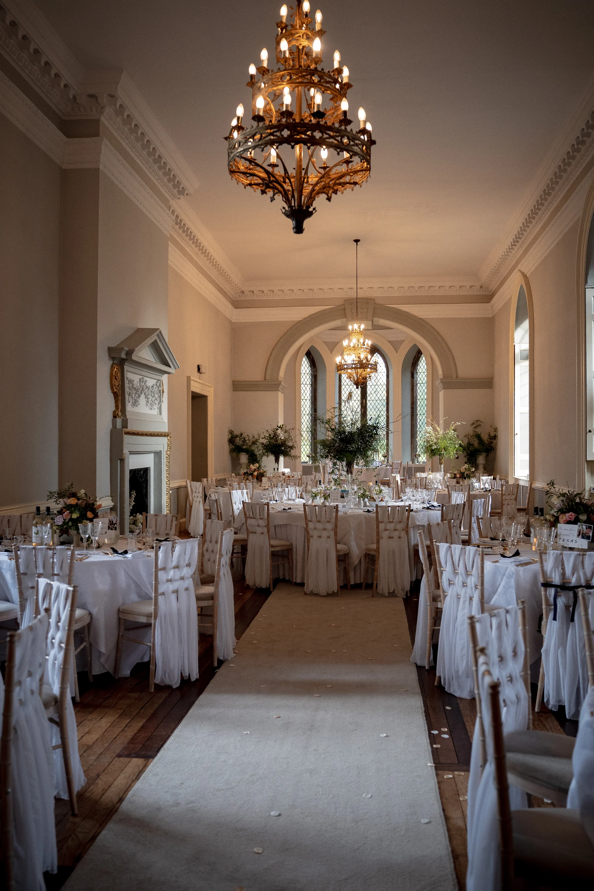 An elegant grand dining room in a stately home is arranged for a wedding reception with round tables featuring white linens and tall chandeliers hanging from the high ceiling.