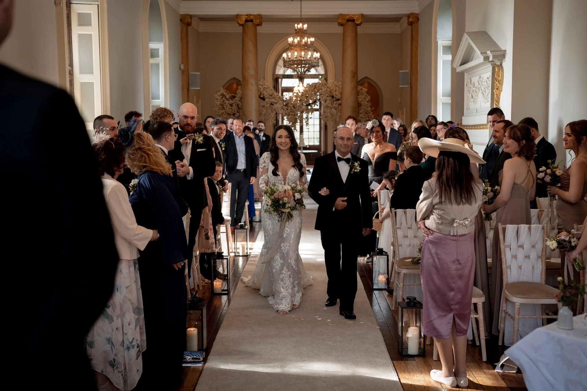A smiling bride holding a bouquet walks down the aisle on her father's arm inside a grand country house venue lined with seated guests.
