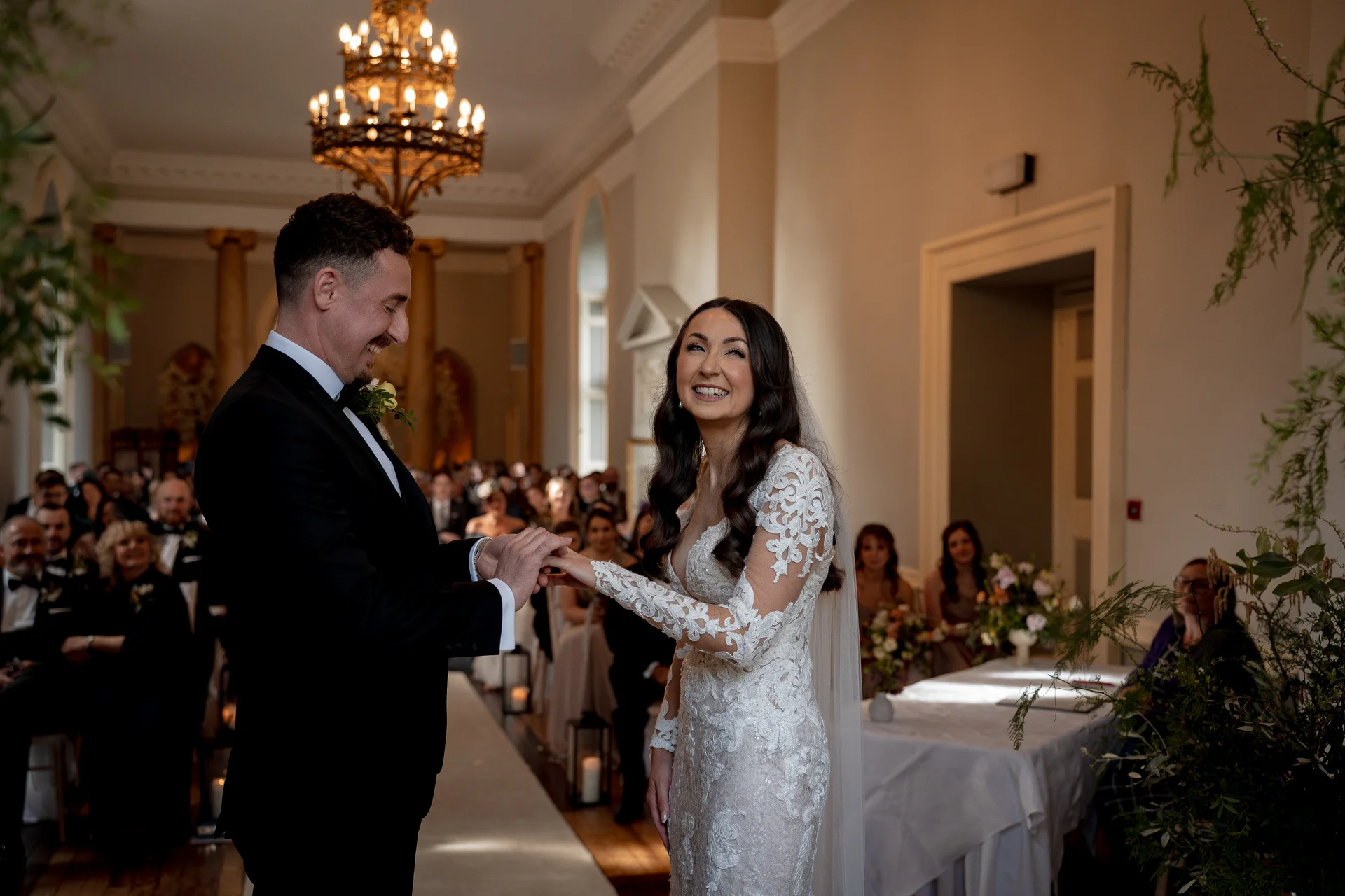 A newlywed groom smiles warmly as he slips a ring onto the laughing bride's finger during their elegant indoor ceremony.