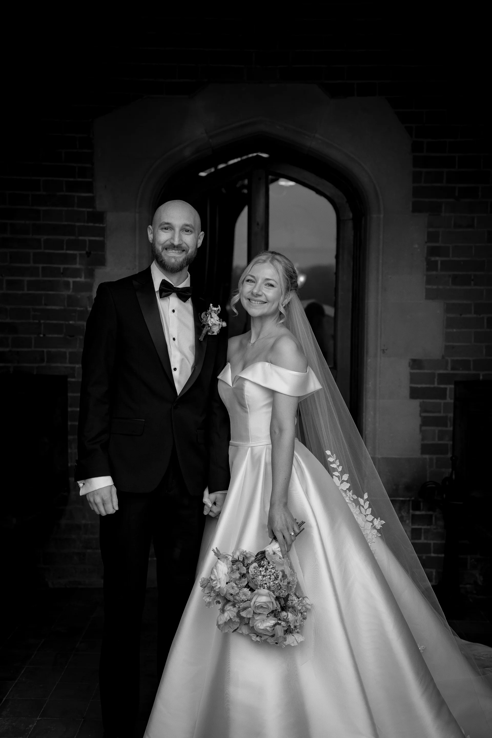 A newlywed couple poses smiling in a black and white portrait in front of a stone doorway, the bride wearing an off-the-shoulder satin gown and holding a bouquet while the groom wears a classic tuxedo.