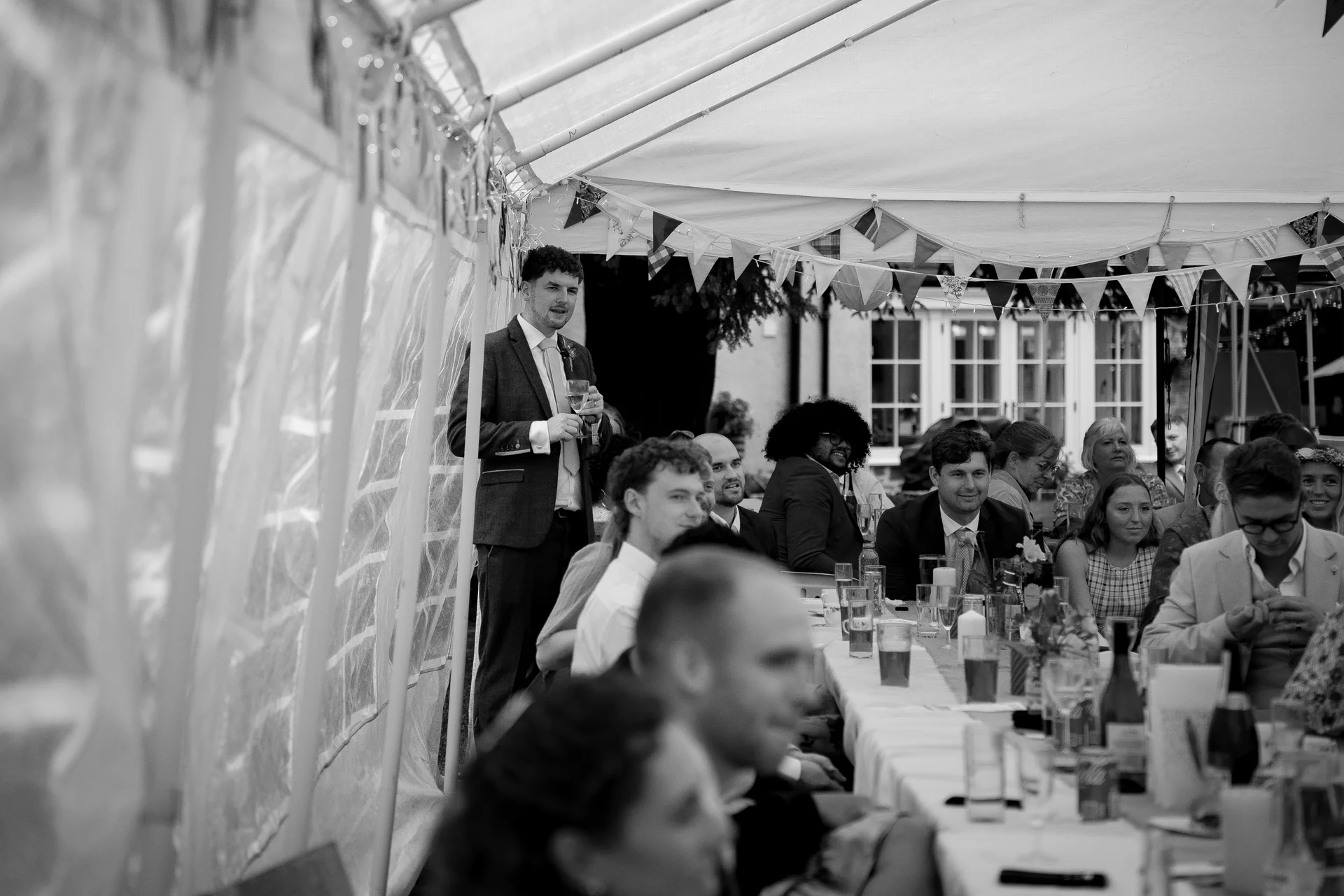A man in a suit stands to deliver a speech during a wedding breakfast inside a marquee decorated with bunting.
