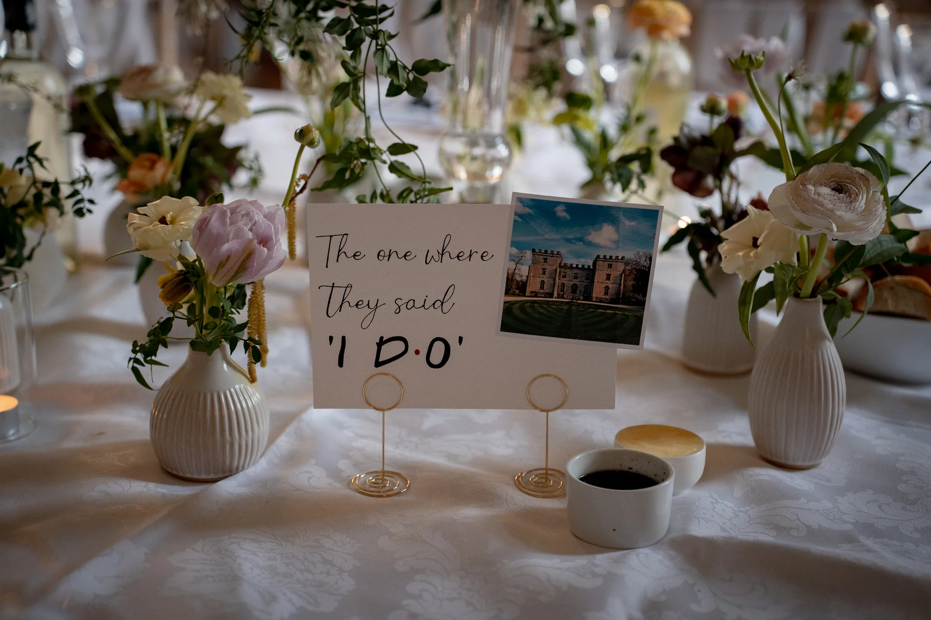 A close-up of a wedding table setting featuring a quirky sign reading "The one where they said I do" alongside delicate bud vases filled with ranunculus and white roses.
